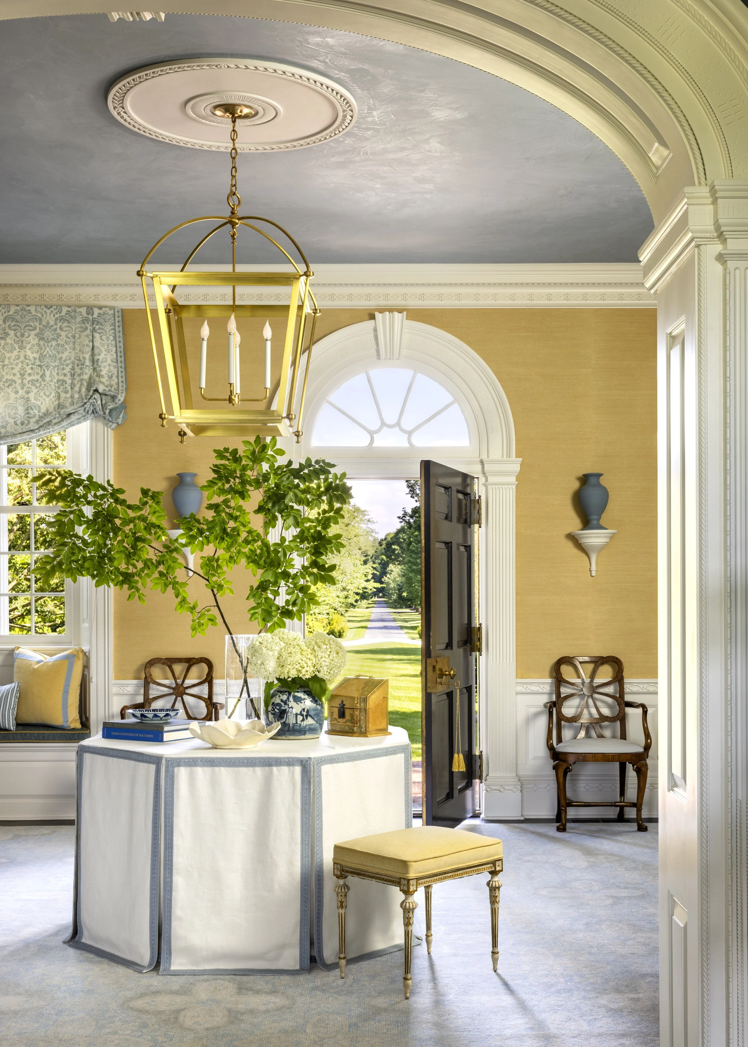 Elegant room with a round table covered with a white tablecloth, decorated with a vase of white hydrangeas, a small wooden chest, and a bowl. The room features a large window with a half-moon top, an open dark wooden door, a brass chandelier with can