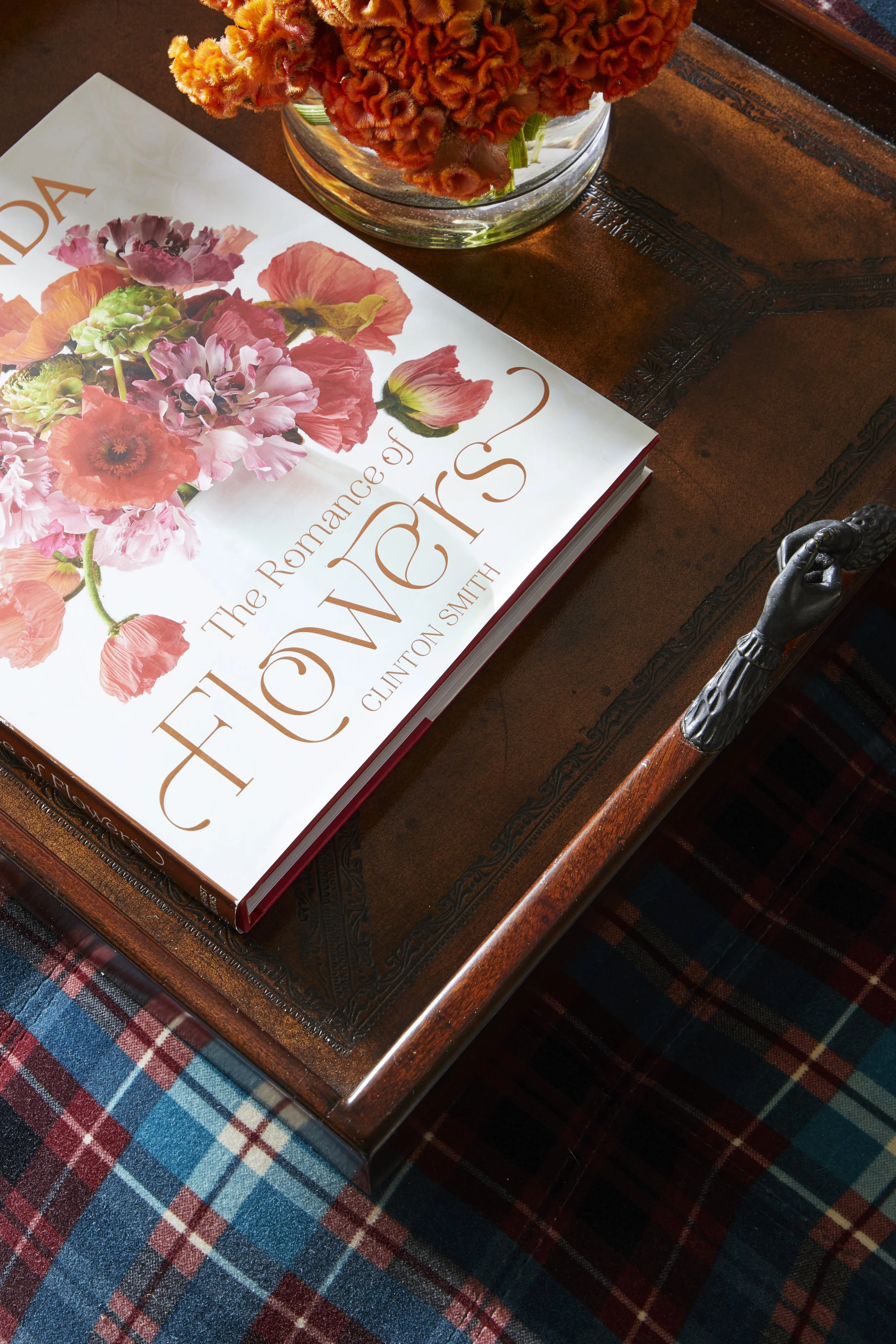 Close-up of a book titled "The Romance of Flowers" by Clinton Smith, resting on an ornate wooden table with metal accents. The table is placed on a plaid patterned carpet with shades of red, blue, and turquoise. There is a glass bowl with orange flow