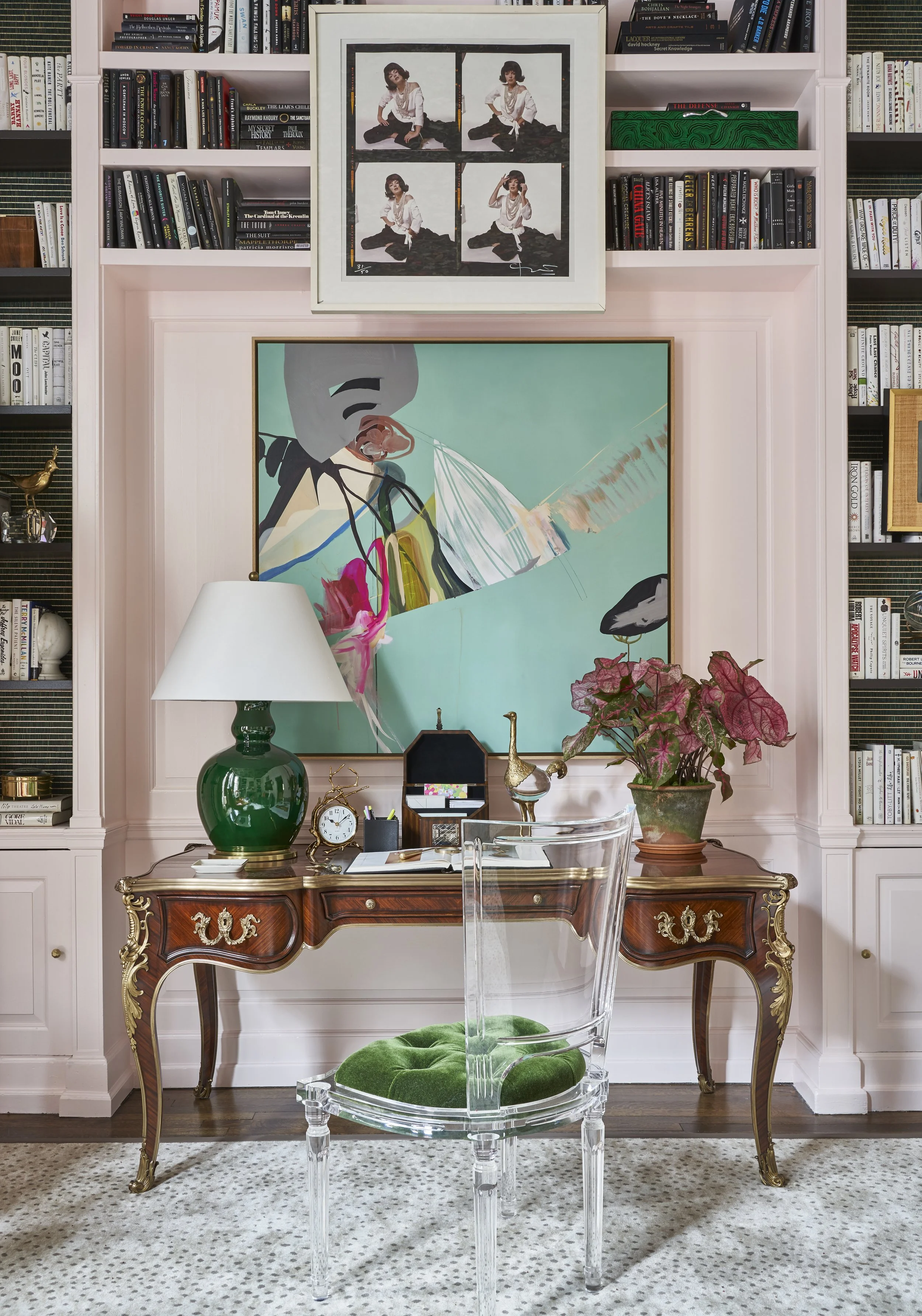 A home office with pink paneled walls and a built-in white bookshelf filled with books. A large colorful abstract painting hangs above a vintage wooden desk with gold accents. On the desk, there is a green glass table lamp, a clock, a small jewelry b