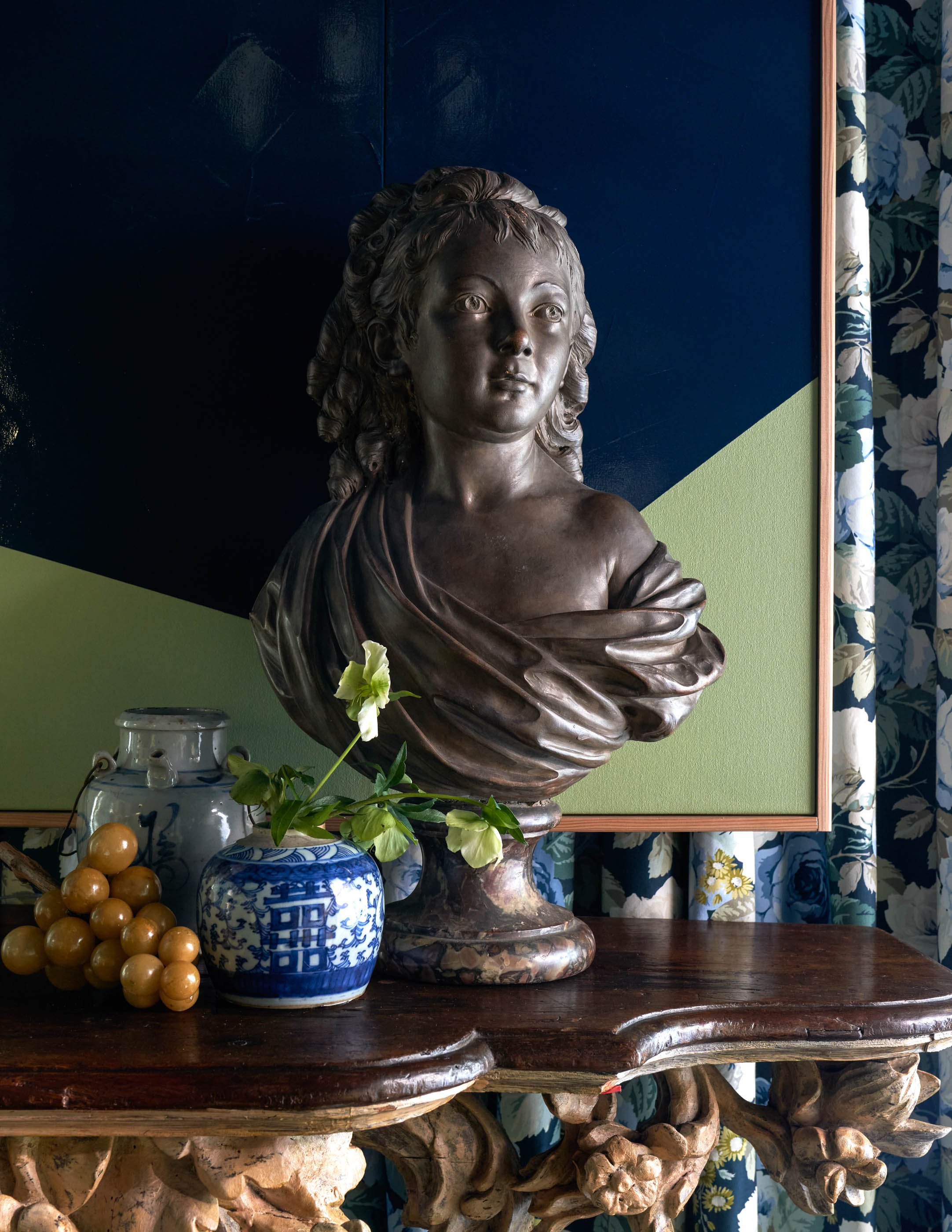 A bronze bust of a woman on a marble pedestal. In front of it, there are blue and white patterned vases with flowers and a string of golden beads. The background features a geometrically painted wall and floral curtains.