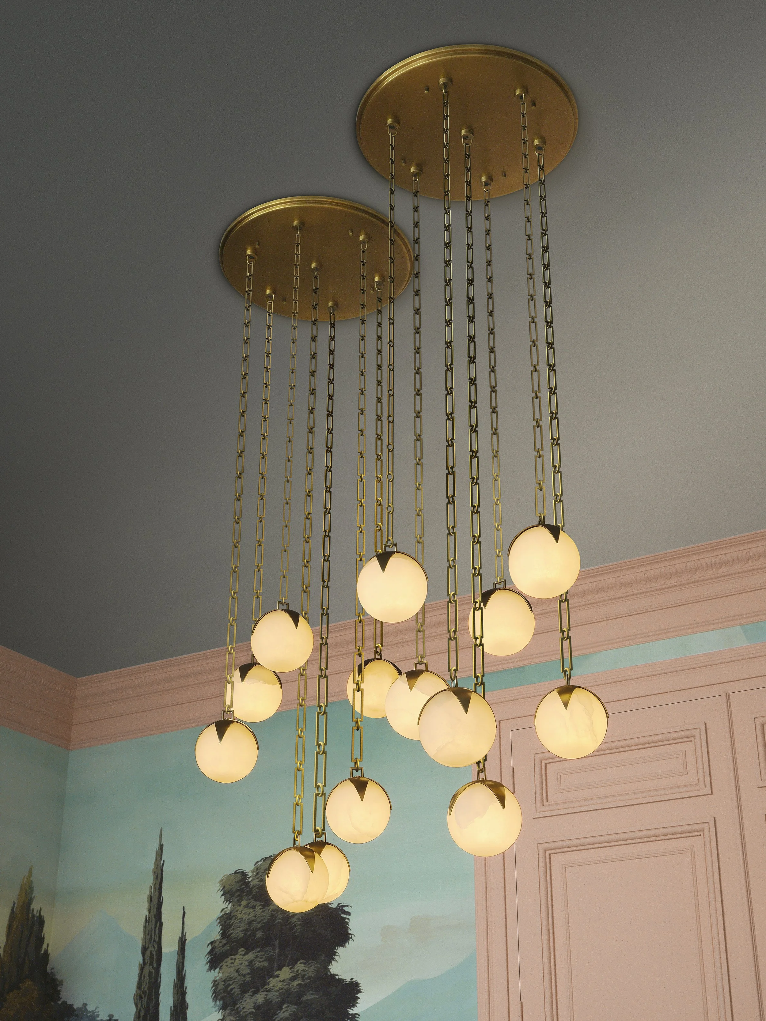 A chandelier with multiple globe-shaped lights hanging from gold chains attached to two gold ceiling mounts in a room with a painted landscape mural and pastel pink and teal walls.