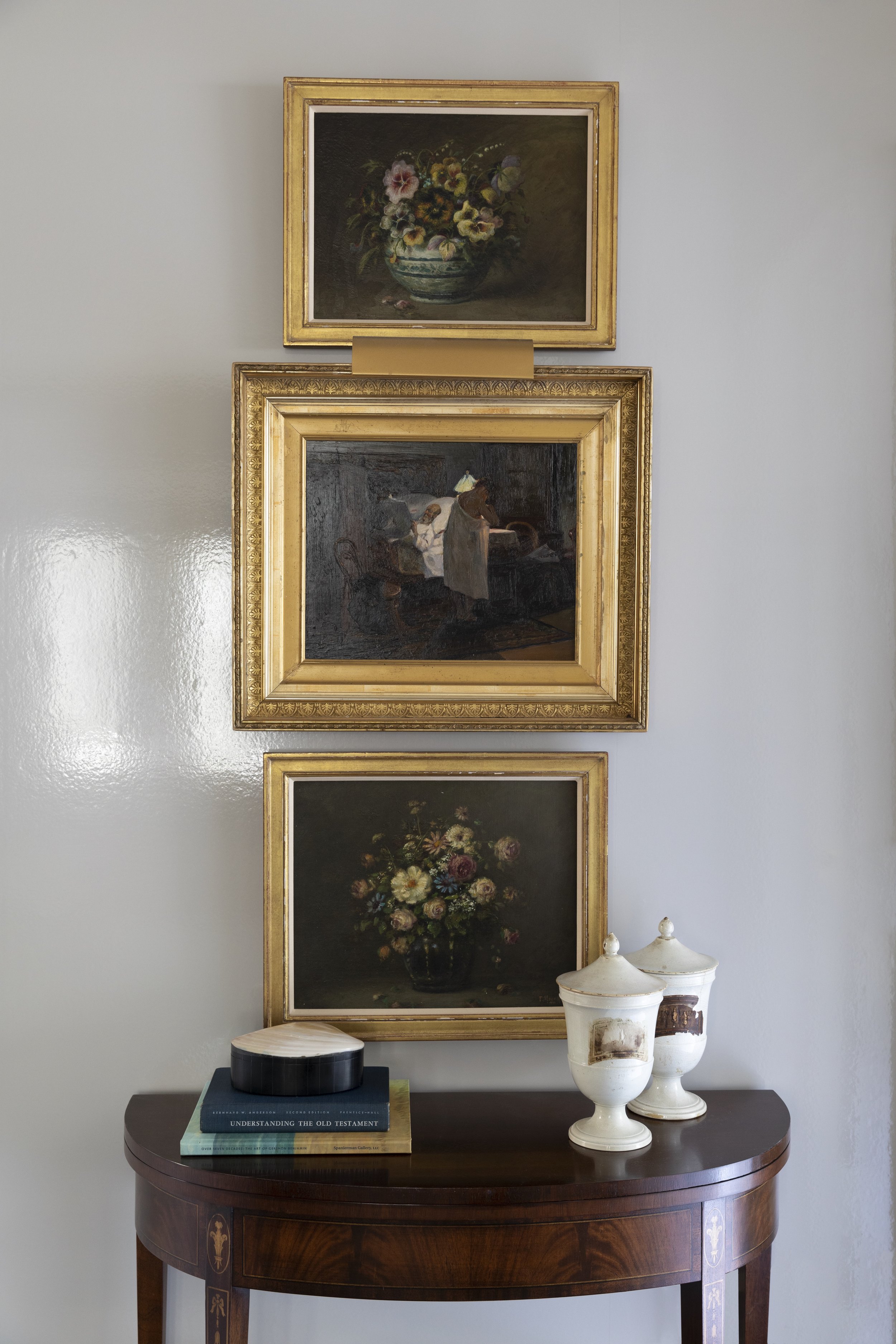 A wooden antique side table with decorative inlay, holding a stack of books and three porcelain vases, positioned in front of a light gray wall. Three framed paintings hang above the table, depicting a floral arrangement, a domestic scene with people