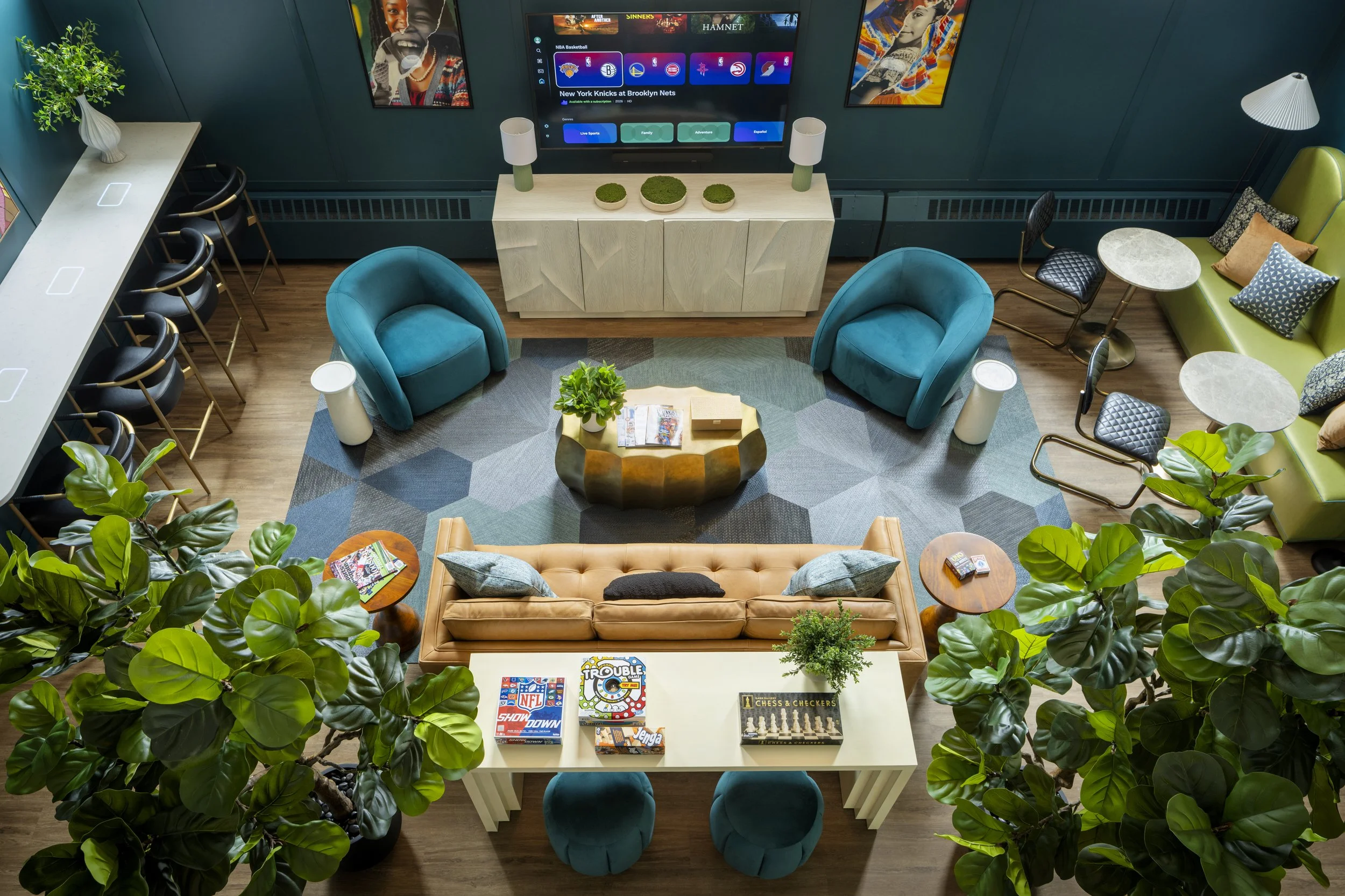 Interior view of a modern living room with green walls, a large television screen, blue armchairs, a green sofa, coffee tables, potted plants, and various board games on a white table.