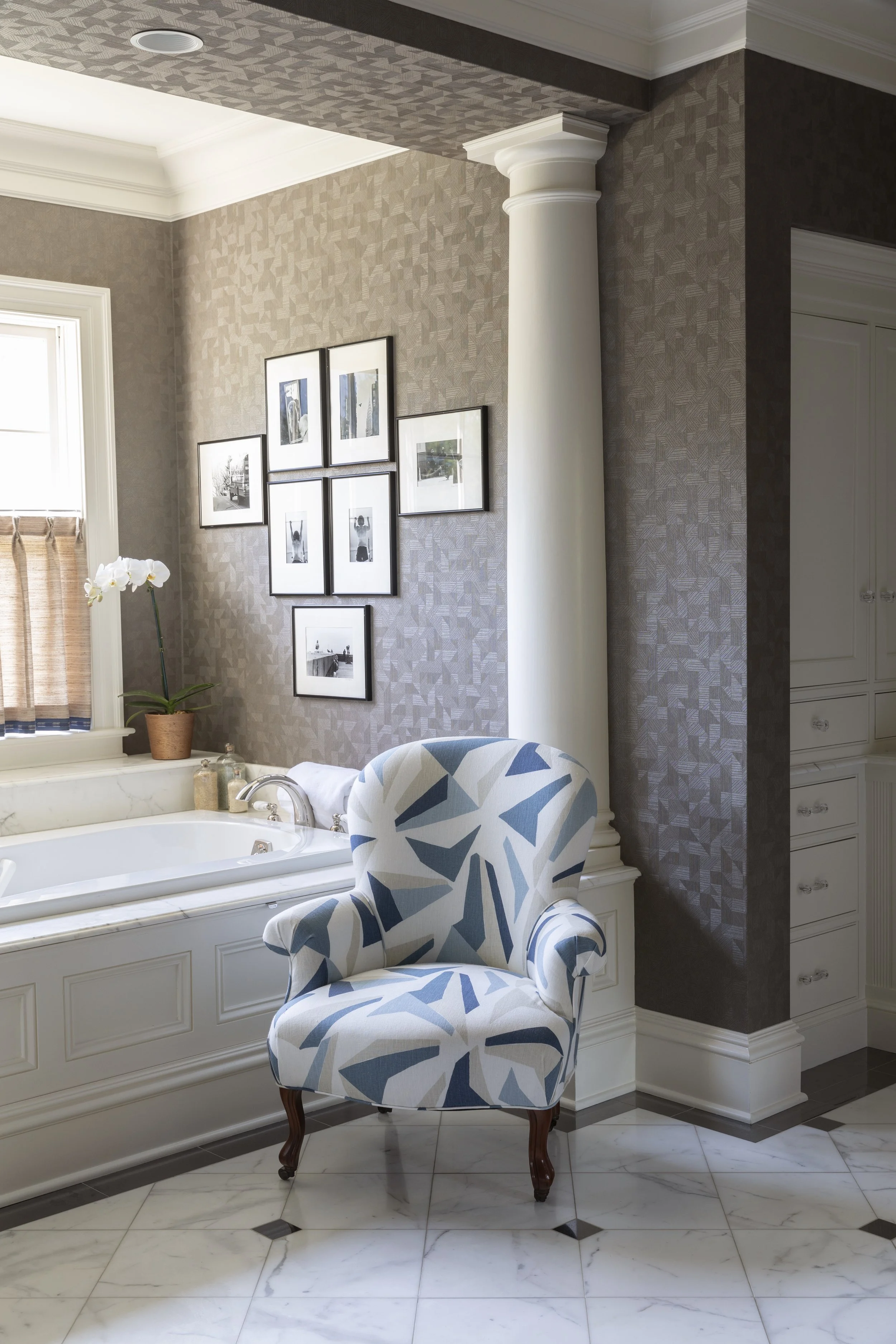A bathroom with a bathtub below a window, decorated with a potted orchid and framed pictures on the wall. A patterned armchair with a blue and gray geometric design is in the foreground. The walls have textured wallpaper, and the flooring features wh