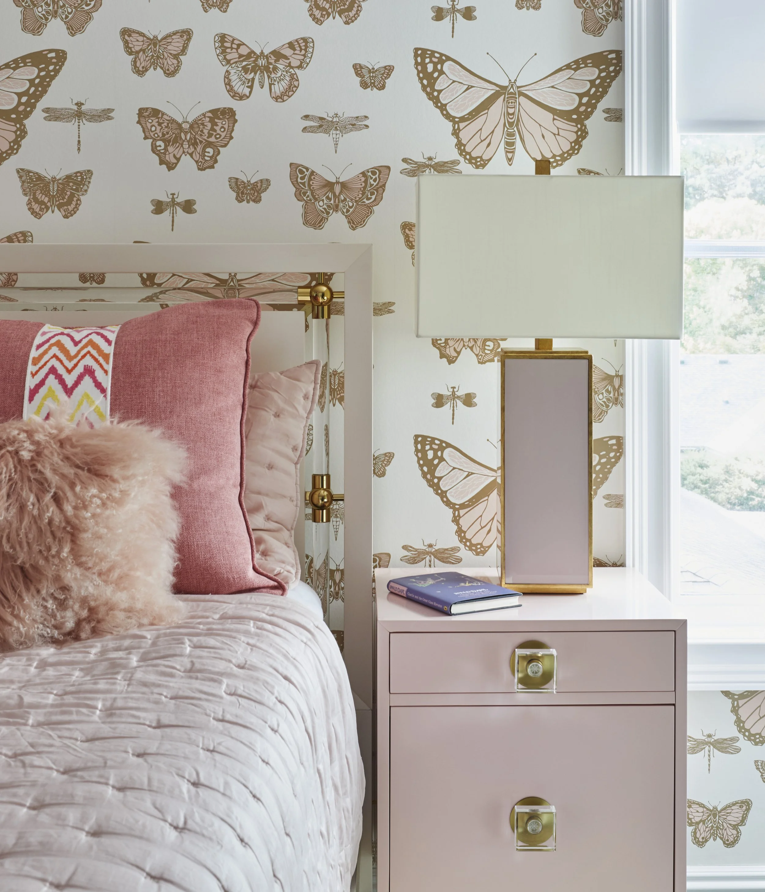 Close-up of a white bedside table with two drawers, one equipped with two glass knobs, placed next to a bed with pink and peach pillows, a fluffy pink throw, and a chevron-patterned pillow. On the table, there is a book titled 'Quiet' by Susan Cain a