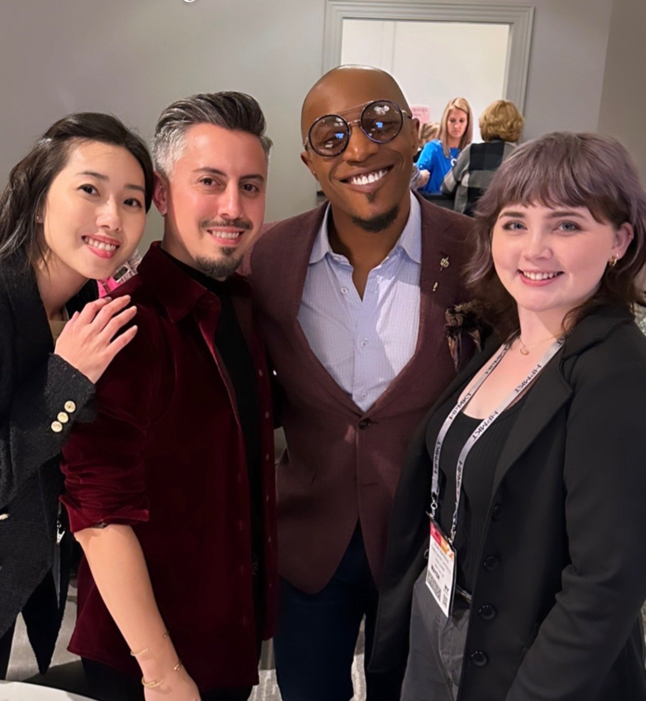 Four people smiling and standing close together in an indoor setting, likely at a conference or event, with two women and two men from diverse backgrounds.