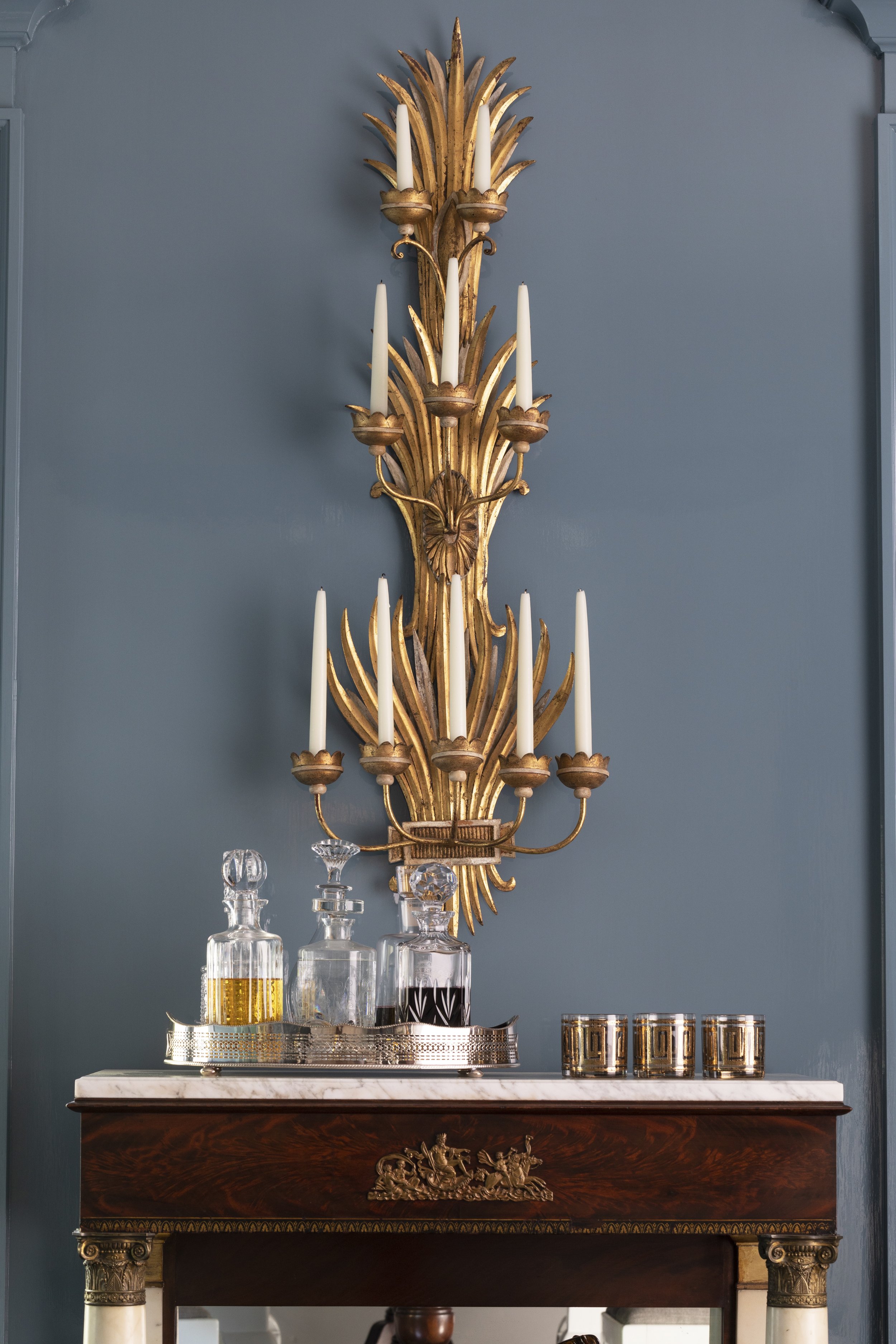 An ornate gold wall sconce with multiple white taper candles on a blue wall, positioned above a marble-topped antique wooden console table with silver decorative elements and glassware.