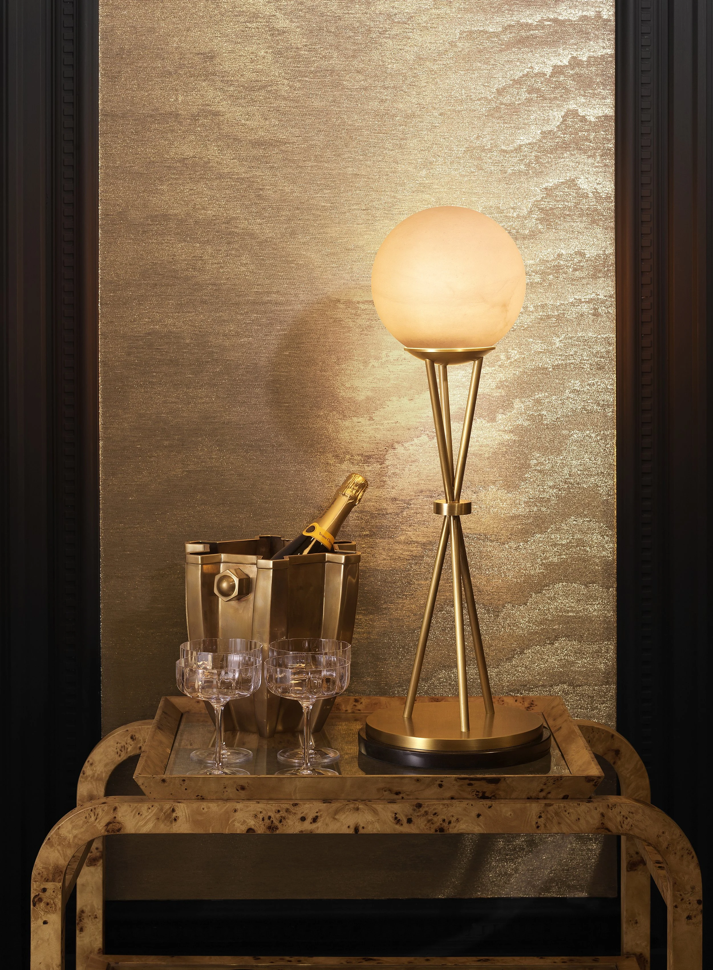 A decorative table setup with a golden floor lamp, a champagne bucket with bottles, three empty glasses, and a wooden tray, set against a textured gold and dark wood framed background.