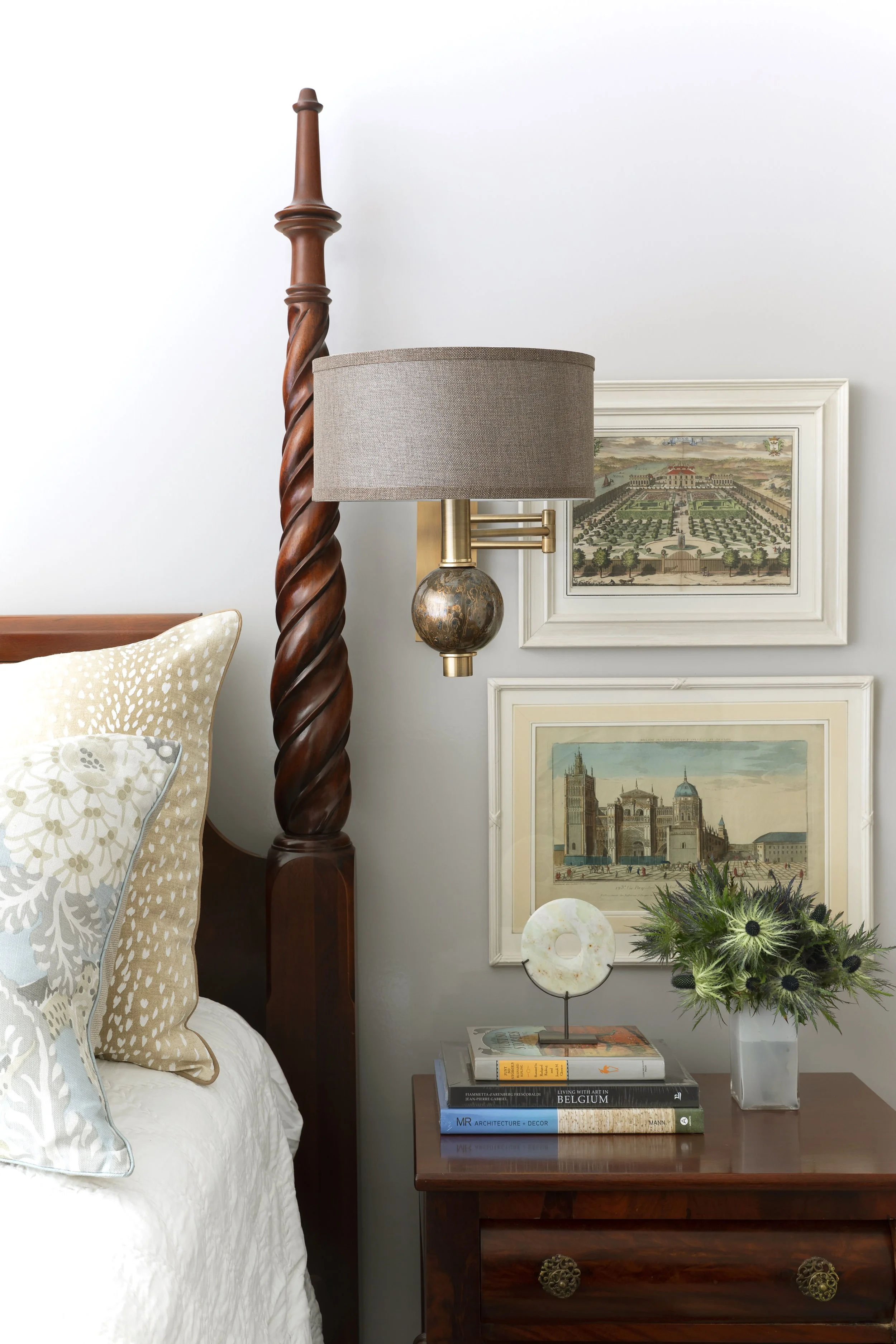 A wooden bedside table with an ornate drawer knob, next to a bed with beige pillows and a floral pillow. Above the table, a modern table lamp with a beige shade, modern brass arm, and a spherical base in a rustic finish. On the wall, there are framed
