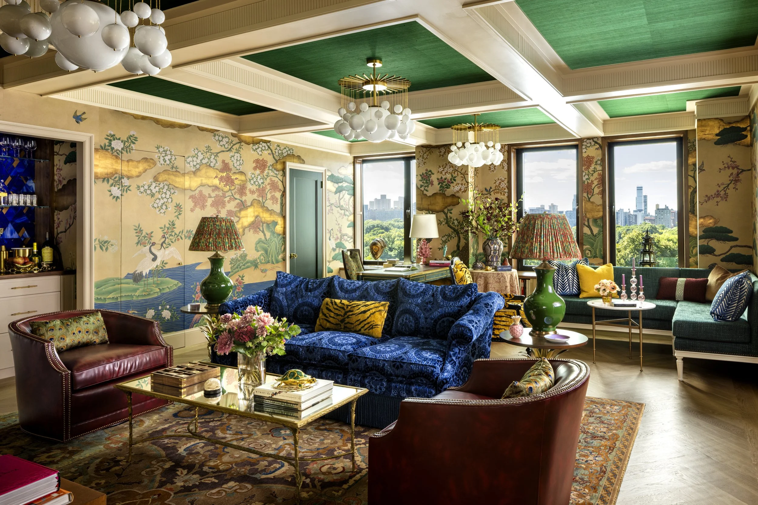 Luxurious living room with large windows, colorful furniture including red, blue, and green sofas, plush pillows, decorated lamps, a coffee table filled with books and decor, lush greenery outside, and elegant chandelier lighting fixtures.