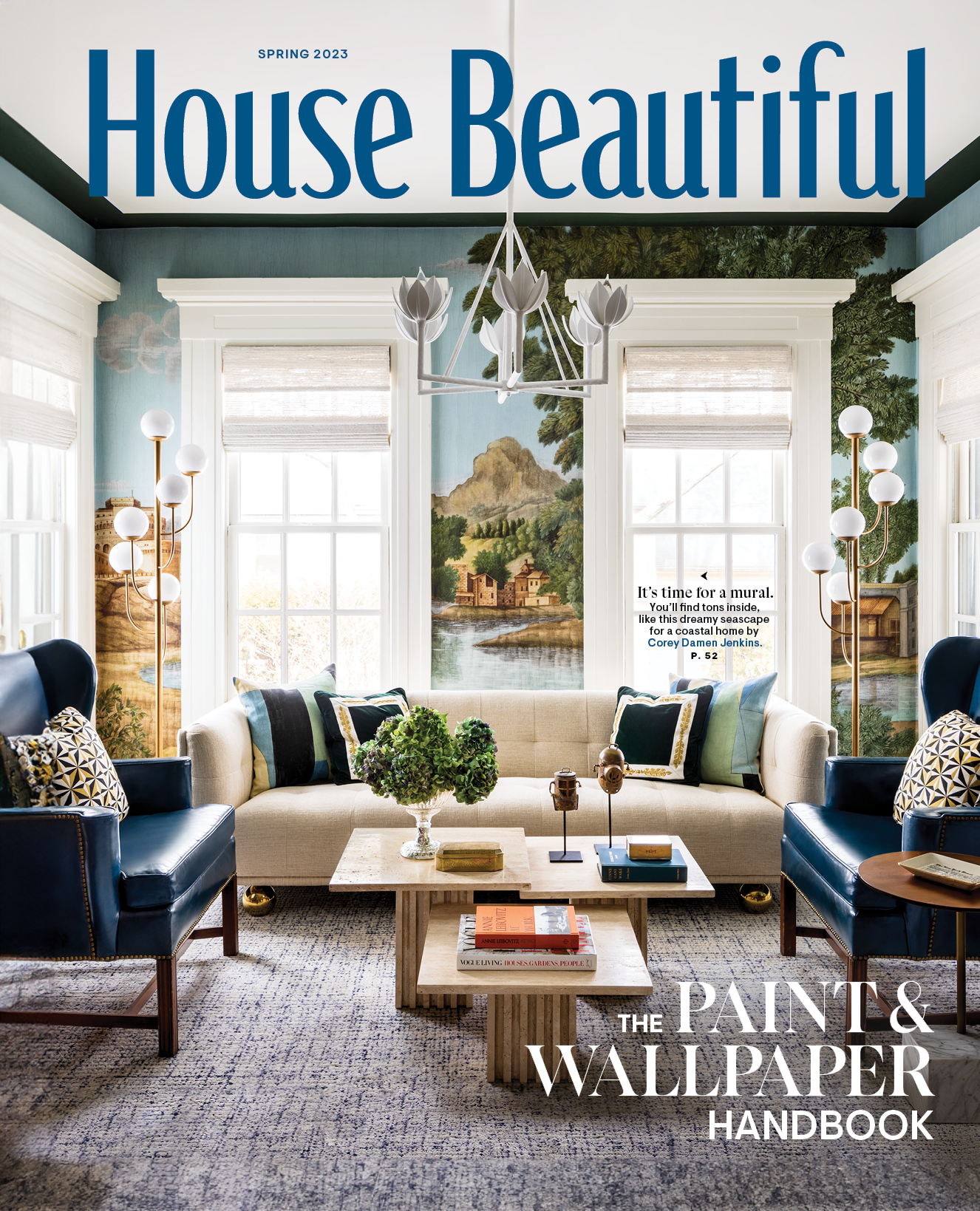 Living room with two white sofas, two navy armchairs, a wooden coffee table, a side table, and decorative pillows. Behind the sofas are three large windows with blinds, and a mural of a coastal landscape with houses, trees, and mountains. A chandelier hangs from the ceiling, and tall floor lamps are on either side of the windows. A book titled 'The Paint & Wallpaper Handbook' is on the coffee table.