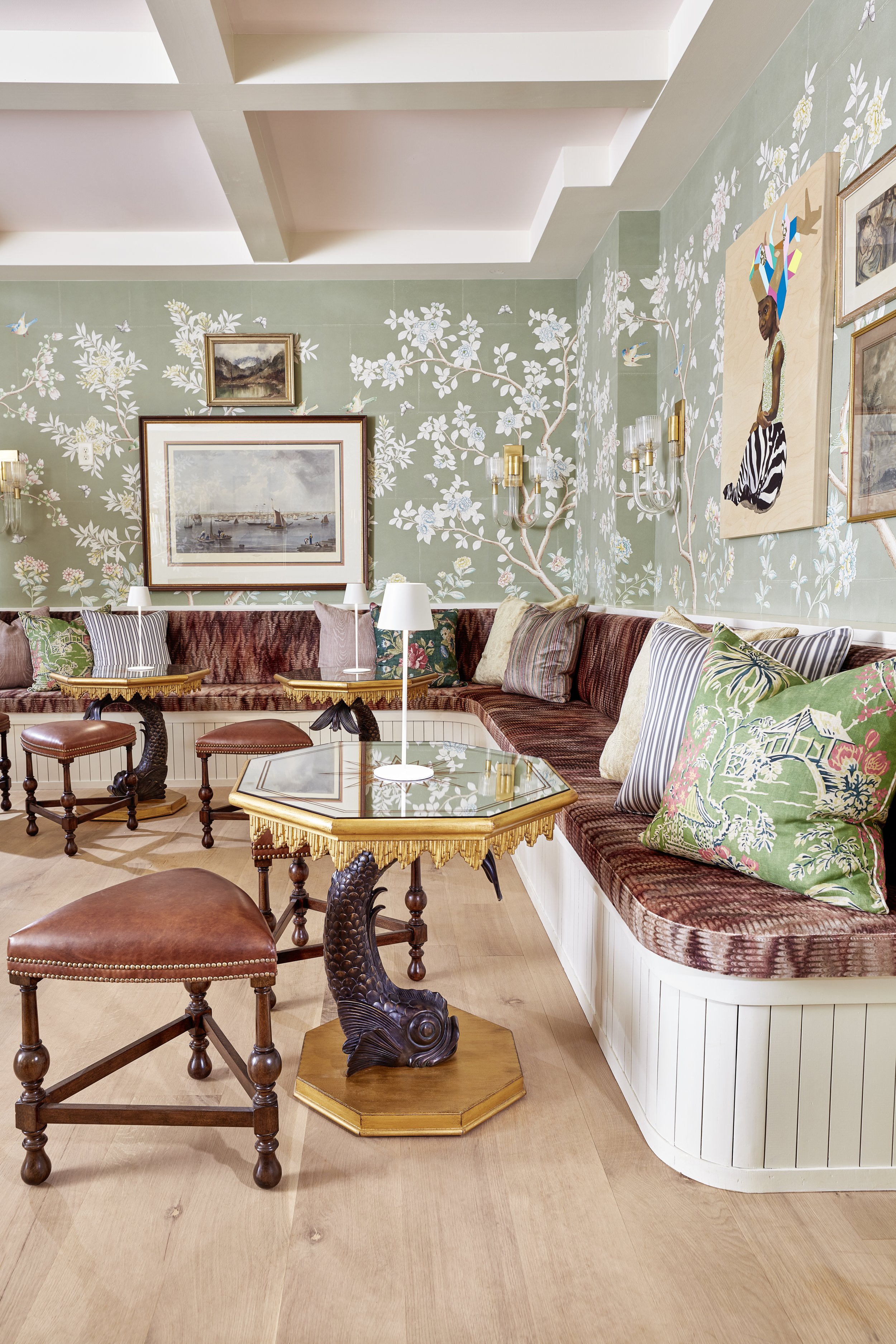 Living room with green floral wallpaper, framed paintings, brass wall sconces, a long maroon upholstered sofa with multicolored cushions, a glass-topped coffee table with a carved wooden base, and various chairs, including a carved fish-shaped wooden