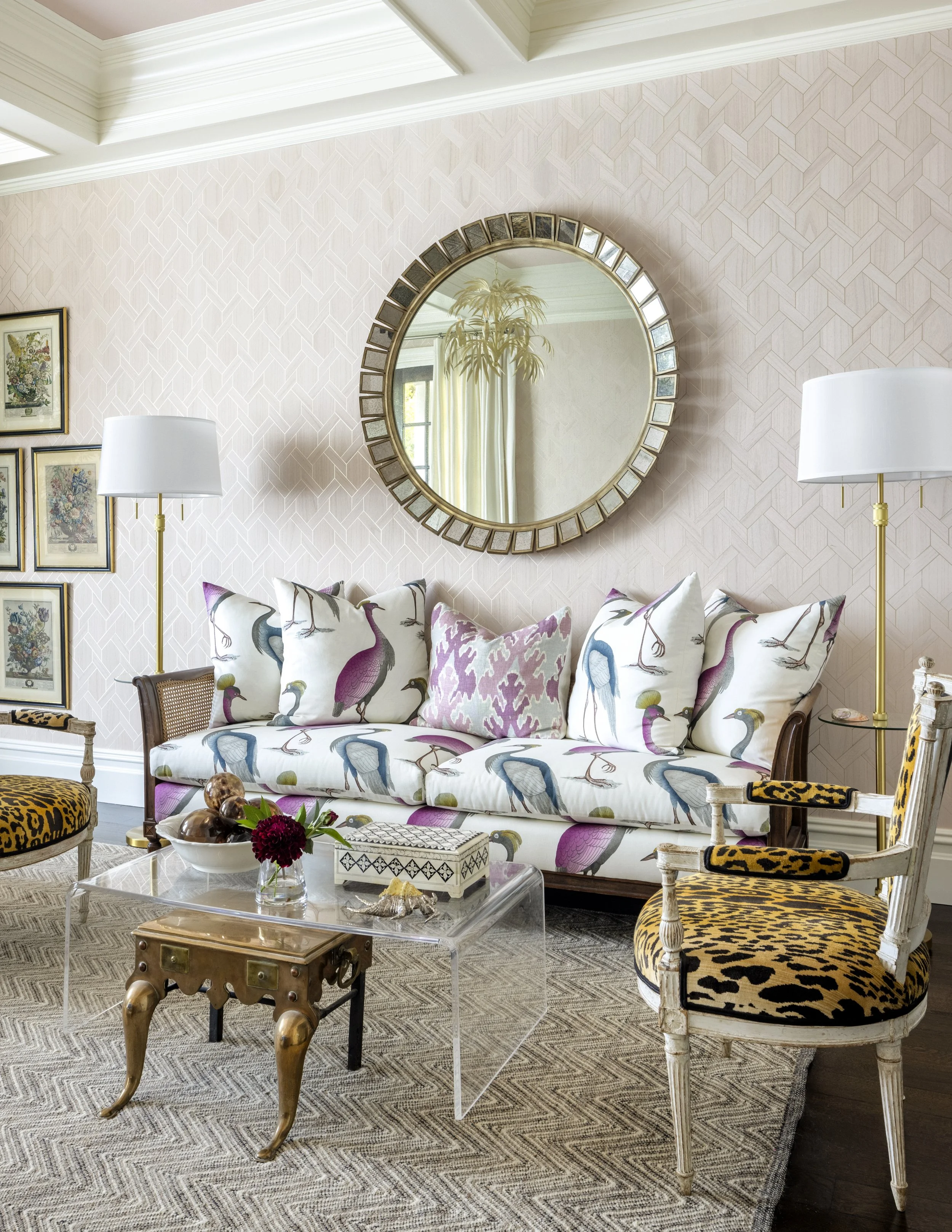 A living room with a patterned pink wall, a round mirror, and a sofa with cushions featuring bird designs. There are two lamps, one on each side of the sofa, and various framed artwork on the wall. A glass coffee table with decorative items and chairs with animal print upholstery are also visible.
