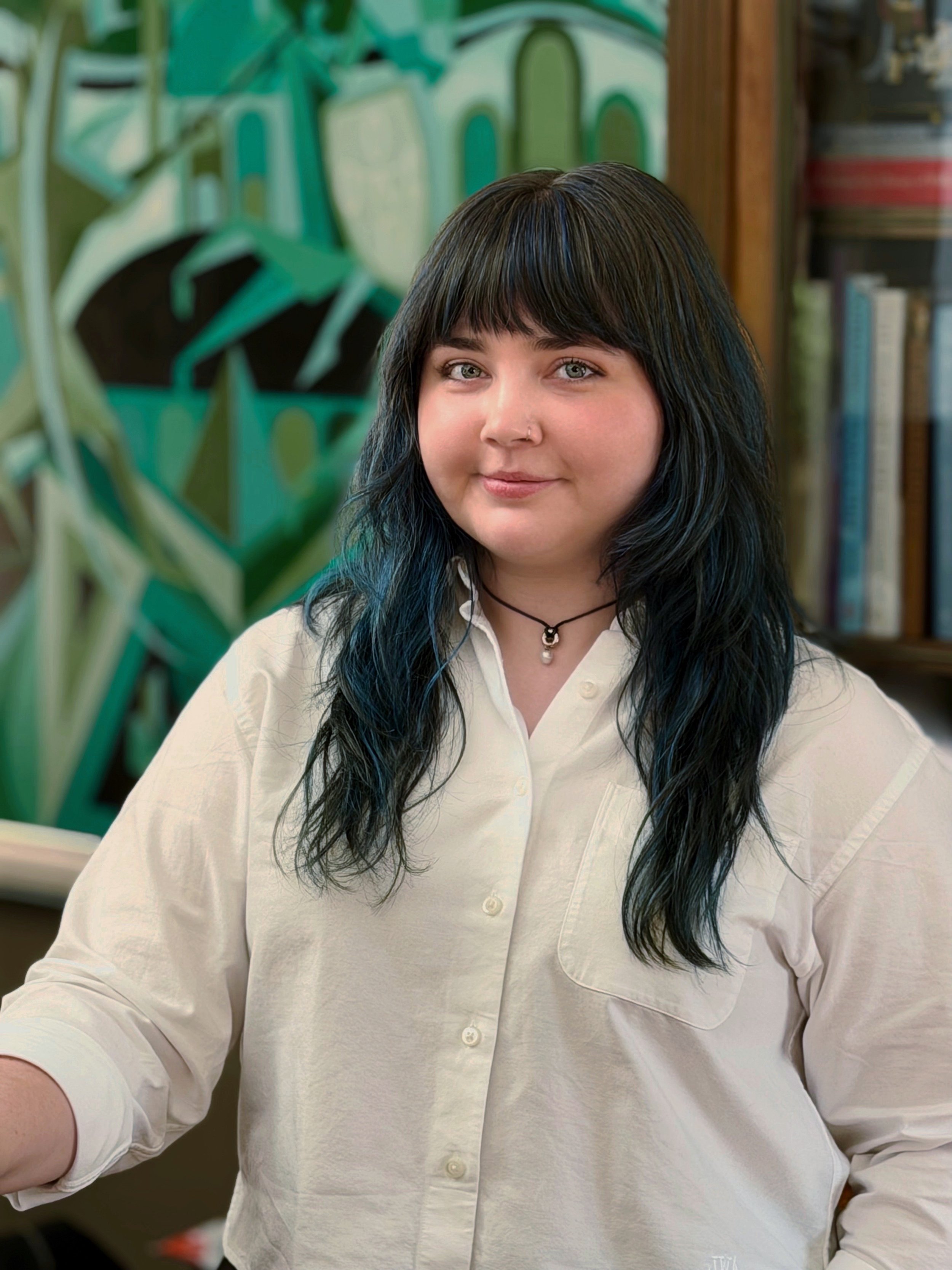 A person with long black hair and light skin wearing a white button-up shirt, sitting in a room with bookshelves and a green plant in the background.