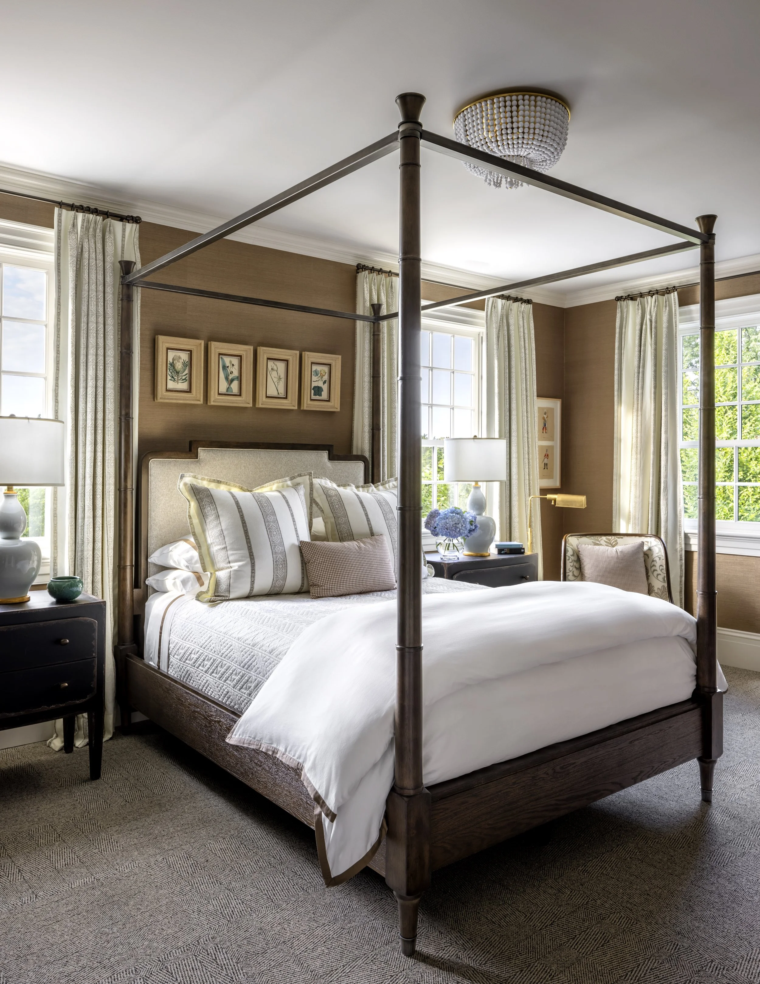 A bedroom with a dark wood four-poster bed, dressed in white and beige striped bedding, flanked by two nightstands with lamps, surrounded by windows with cream curtains, and decorated with framed botanical art.