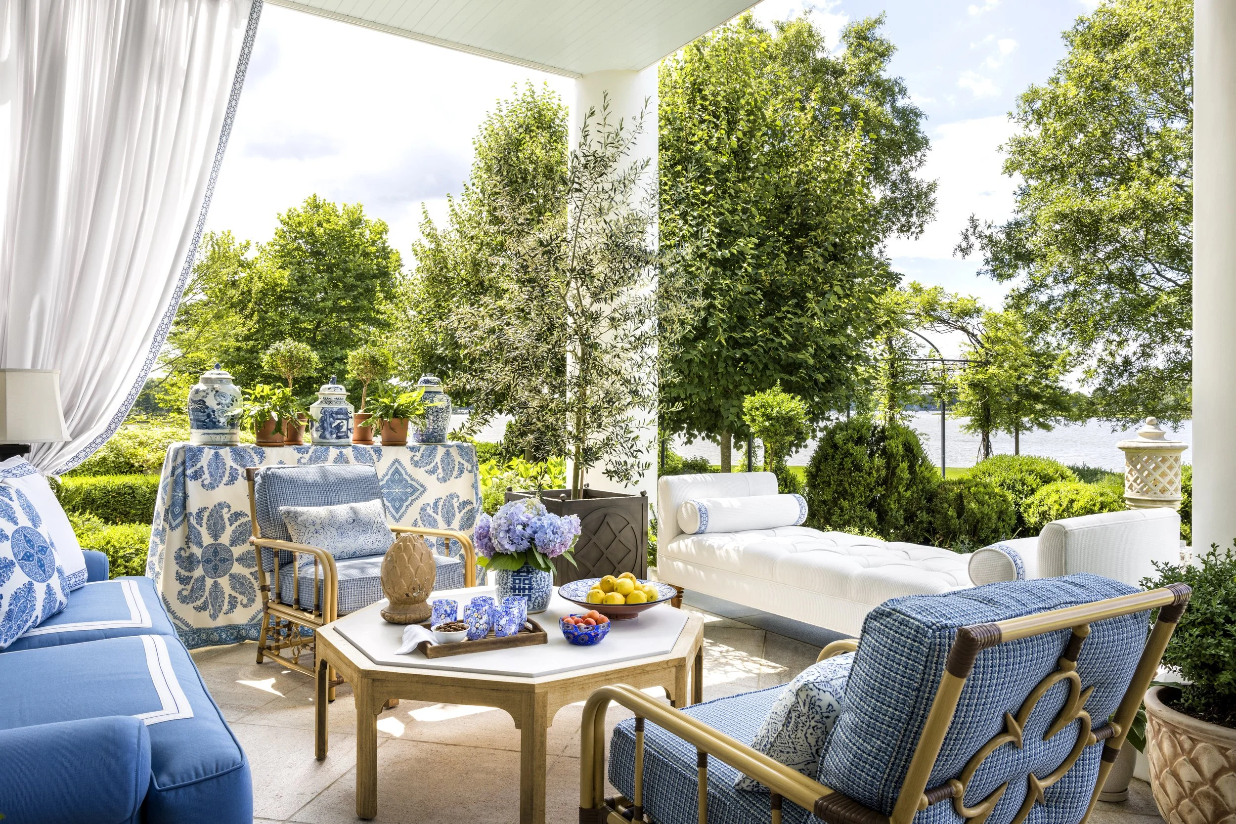 Outdoor patio setup with blue and white patterned furniture, a white chaise lounge, and a table with blue and white ceramics, greenery, trees, and a body of water in the background.
