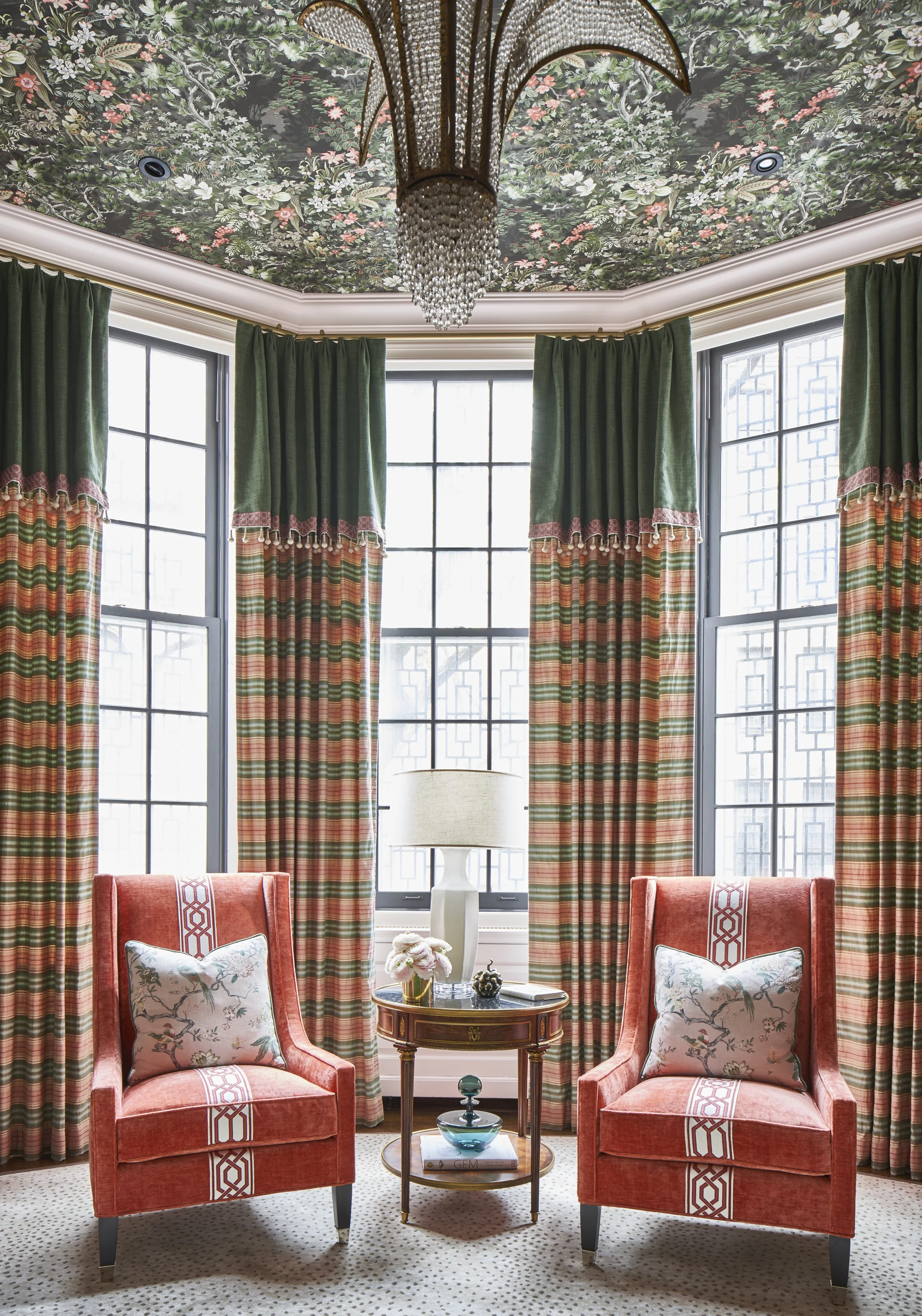 A cozy sitting area with two red velvet armchairs each decorated with a floral-patterned pillow, a round wooden side table with a book and decorative items, large windows with multiple panes, dark green and plaid curtains, a white table lamp, and a f