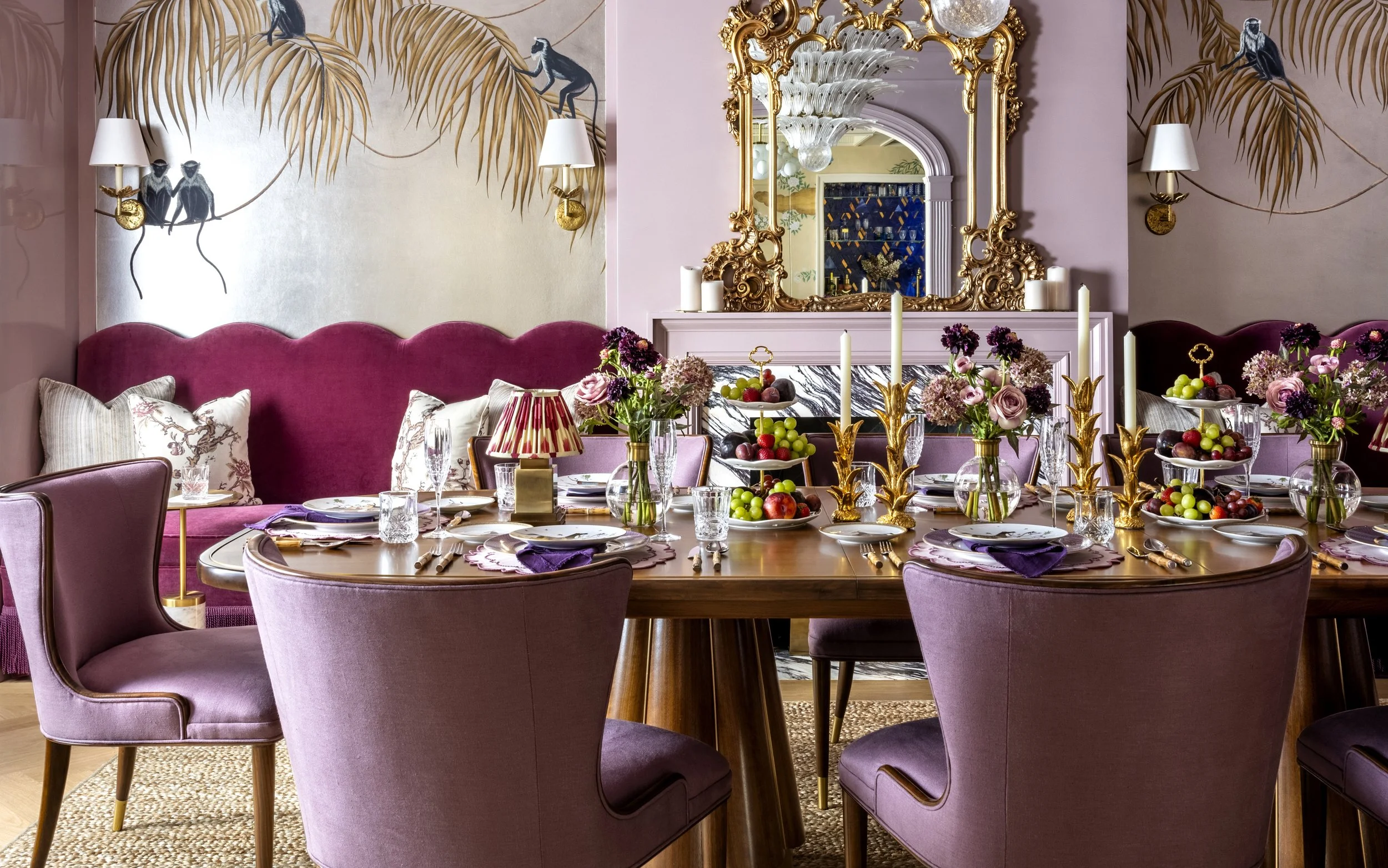 A decorated dining room with pink and purple tones, featuring a round wooden table set with plates, glasses, purple napkins, and floral centerpieces. Pink velvet chairs surround the table, and a large pink velvet sofa is against the wall. The wall ha