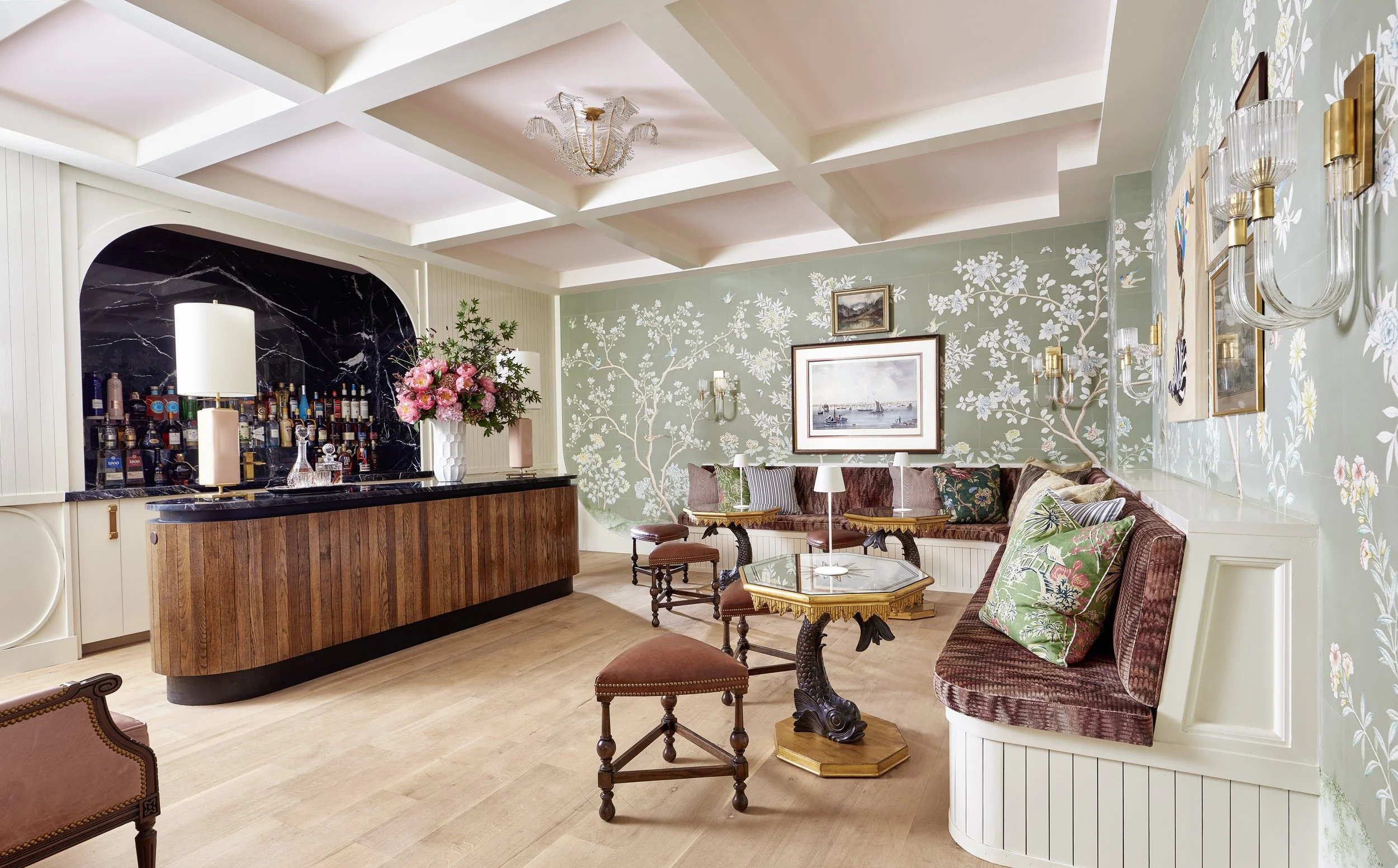 A cozy living room with floral wallpaper, a chandelier, a bar area with bottles, and a sofa with patterned pillows.