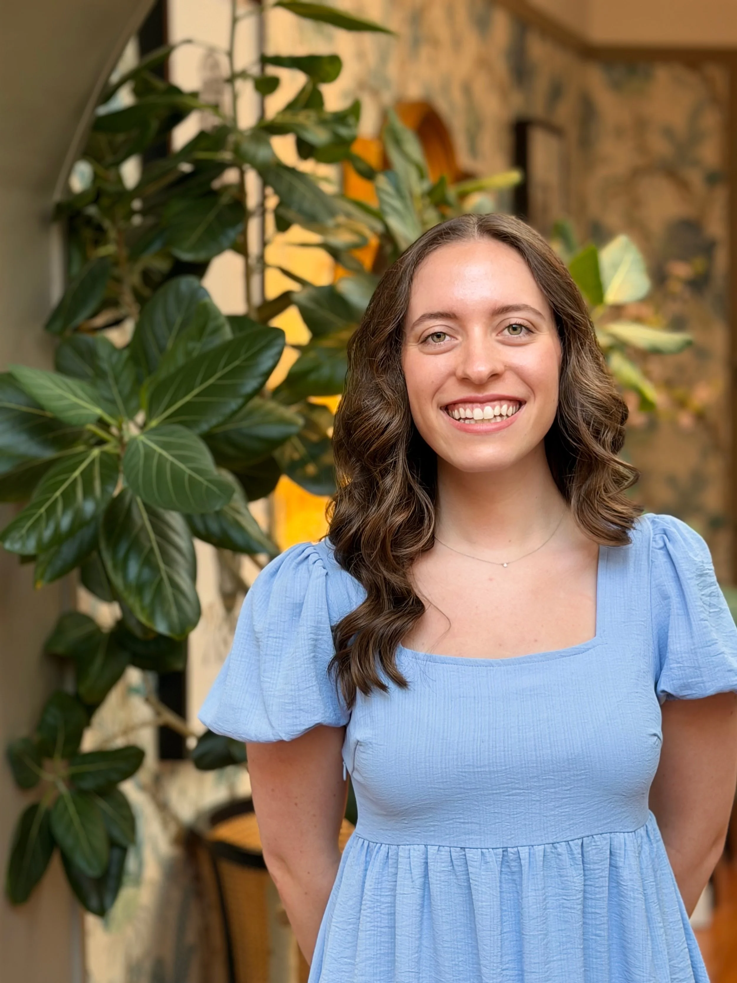 A woman with long, wavy brown hair and a bright smile, wearing a light blue dress, standing indoors near a large houseplant, with a patterned wall in the background.