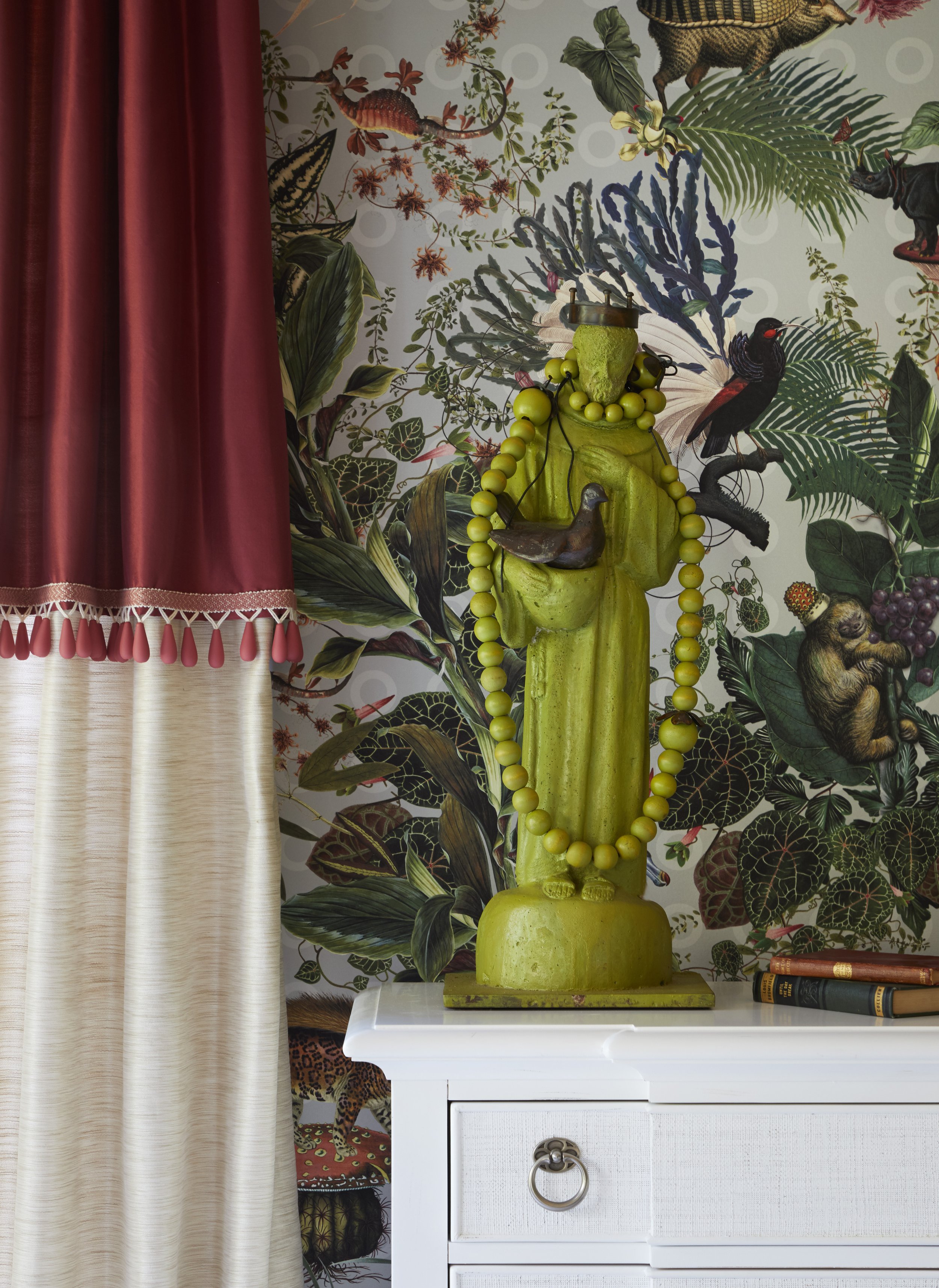 Decorative green sculpture of a figure with a bird on shoulder, adorned with a necklace of yellow beads, placed on a white cabinet. Bright wallpaper with tropical foliage and animals, partially covered by a red curtain with pink tassels on the left a