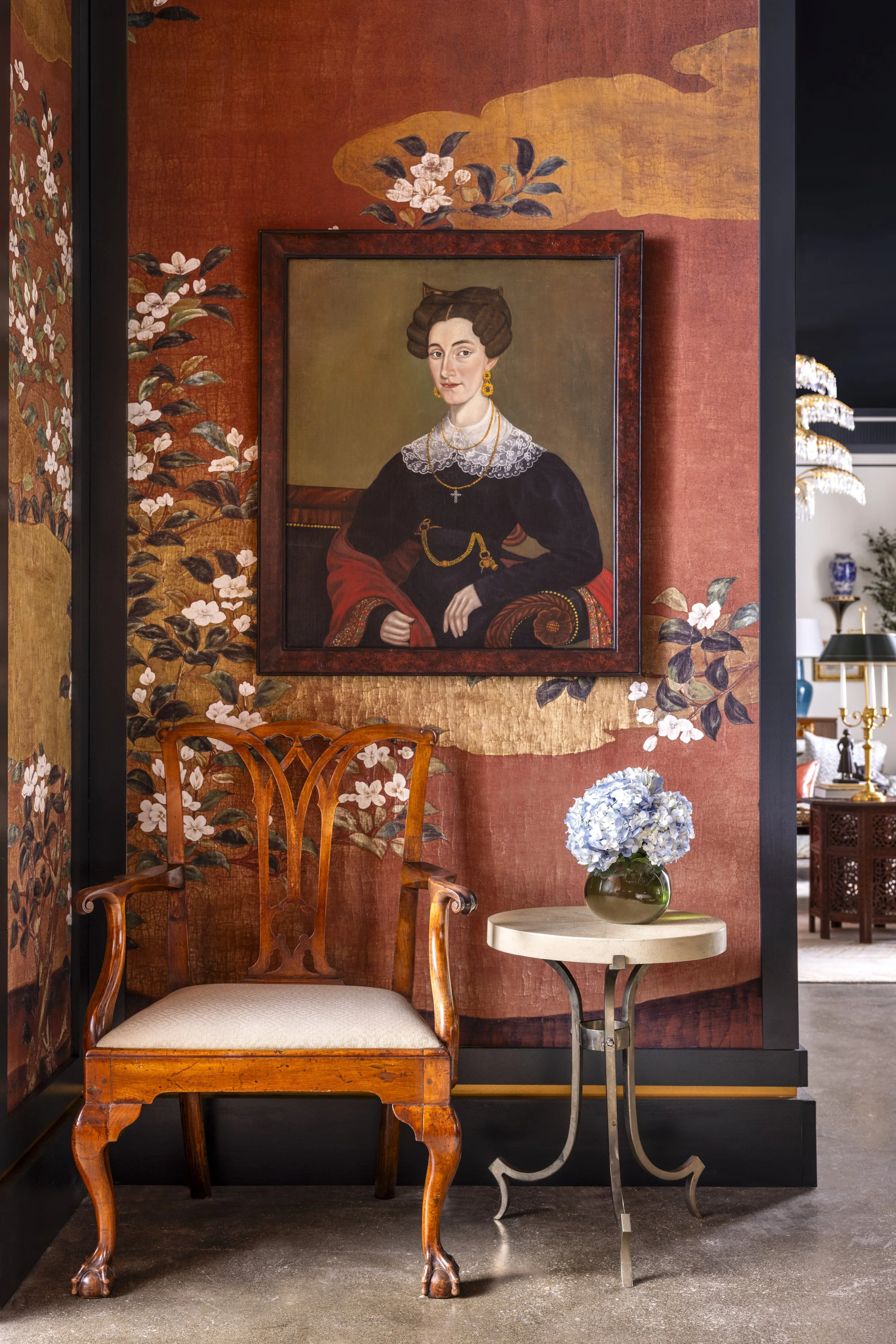 A corner of a room with an antique wooden chair with a cushioned seat, a small round table with a flowering plant in a vase, and a framed portrait of a woman wearing jewelry and a dark dress, hanging on a decorated wall with floral patterns and a bac