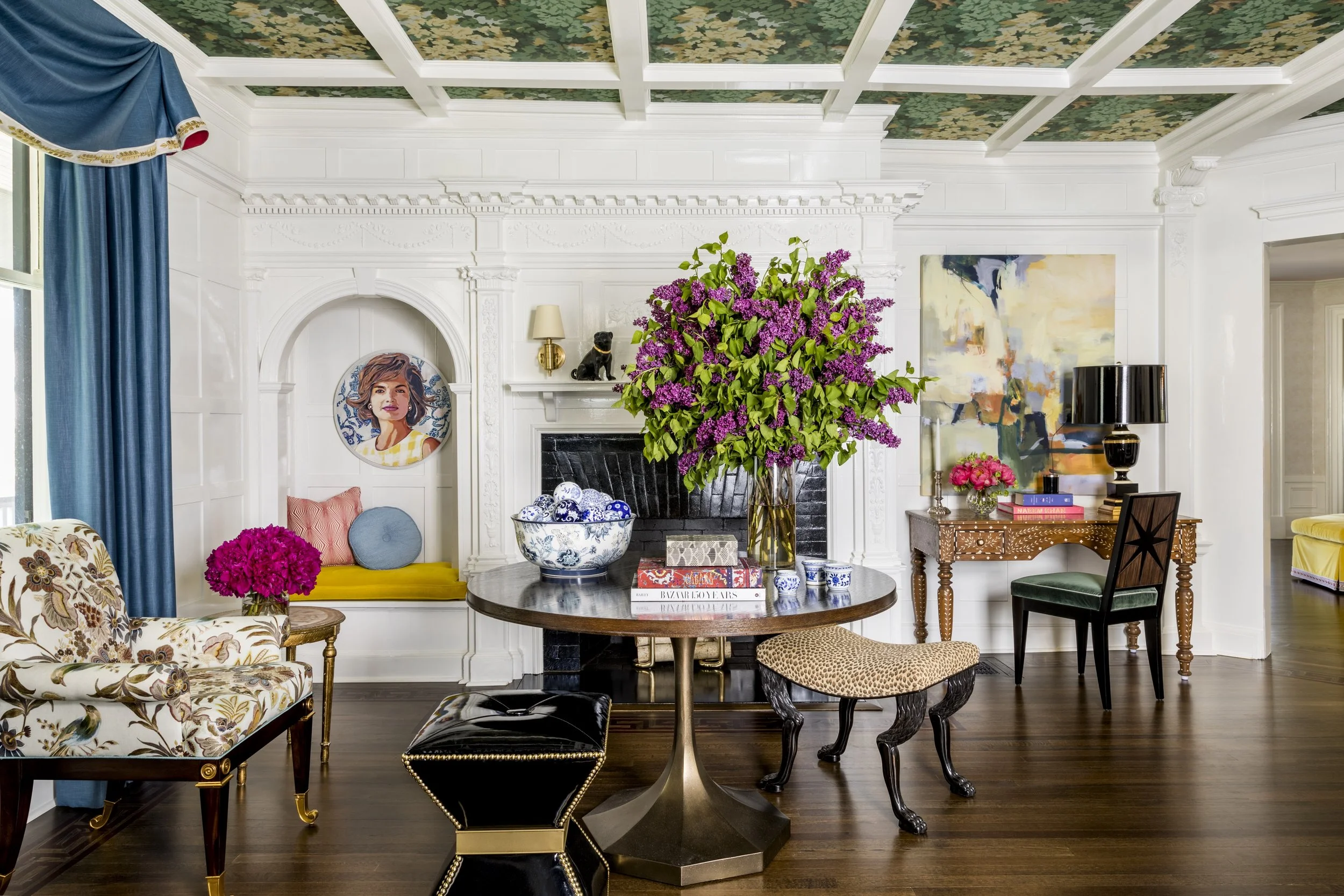 A well-decorated living room with white walls and intricate crown molding. The ceiling has a detailed painted design with green and gold accents. A large vase of purple lilacs and other smaller floral arrangements sit on a coffee table in the center.