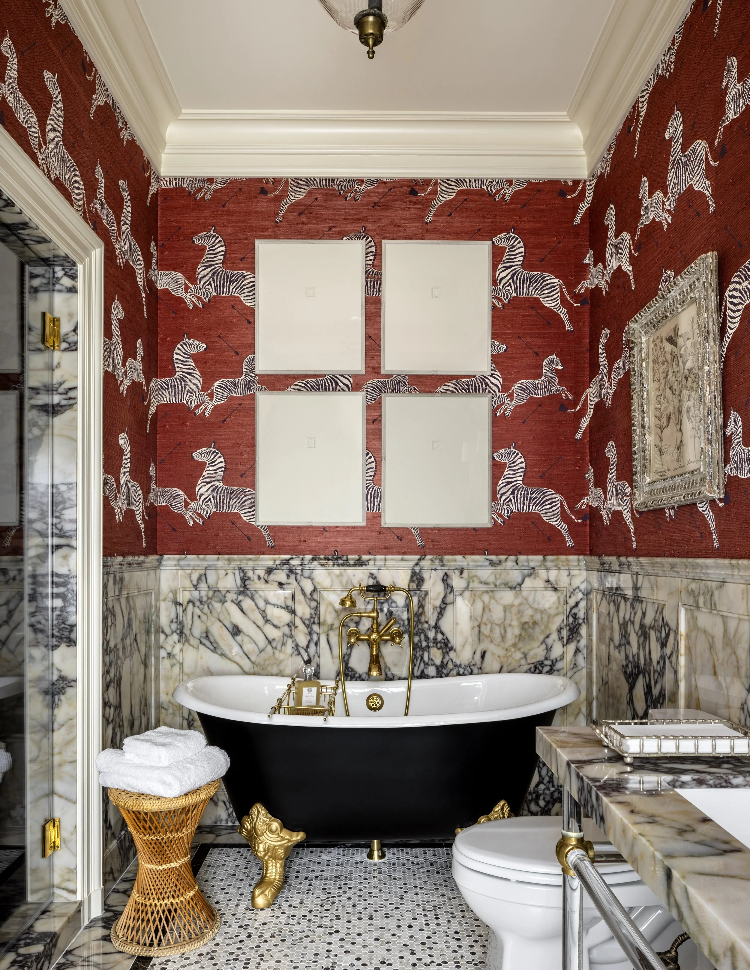 Bathroom with red wallpaper featuring white zebras and black arrows, bathtub with gold clawfoot legs, black and white marble walls, a toilet, a marble countertop, a framed vintage botanical print, a wicker basket with towels, and ceiling crown moldin