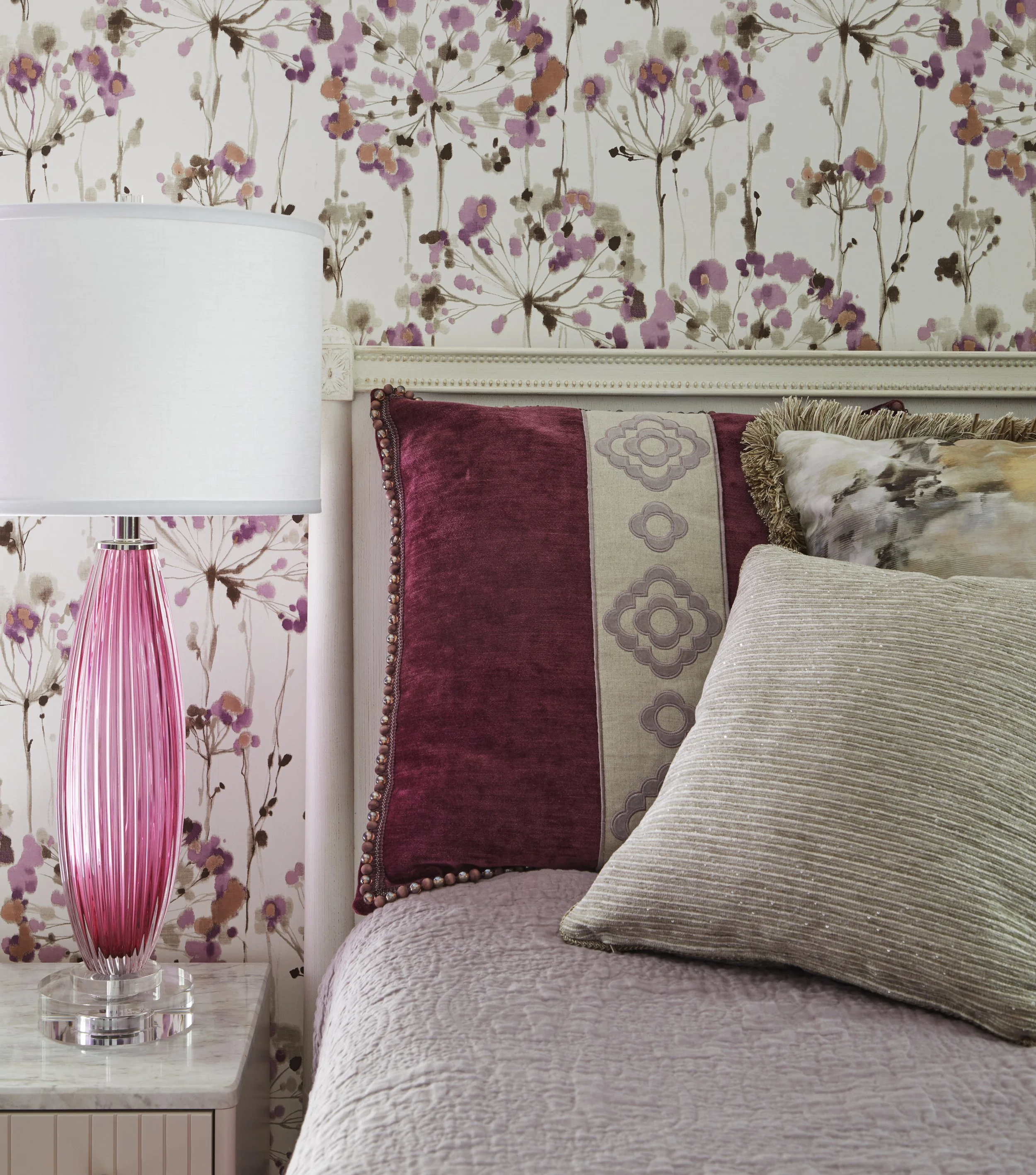 A bedroom scene featuring a bed with cream-colored textured bedding, stacked pillows with various patterns and colors including beige, gold, and maroon, and a floral wallpaper background with pink, purple, brown, and beige flowers. To the left, a whi