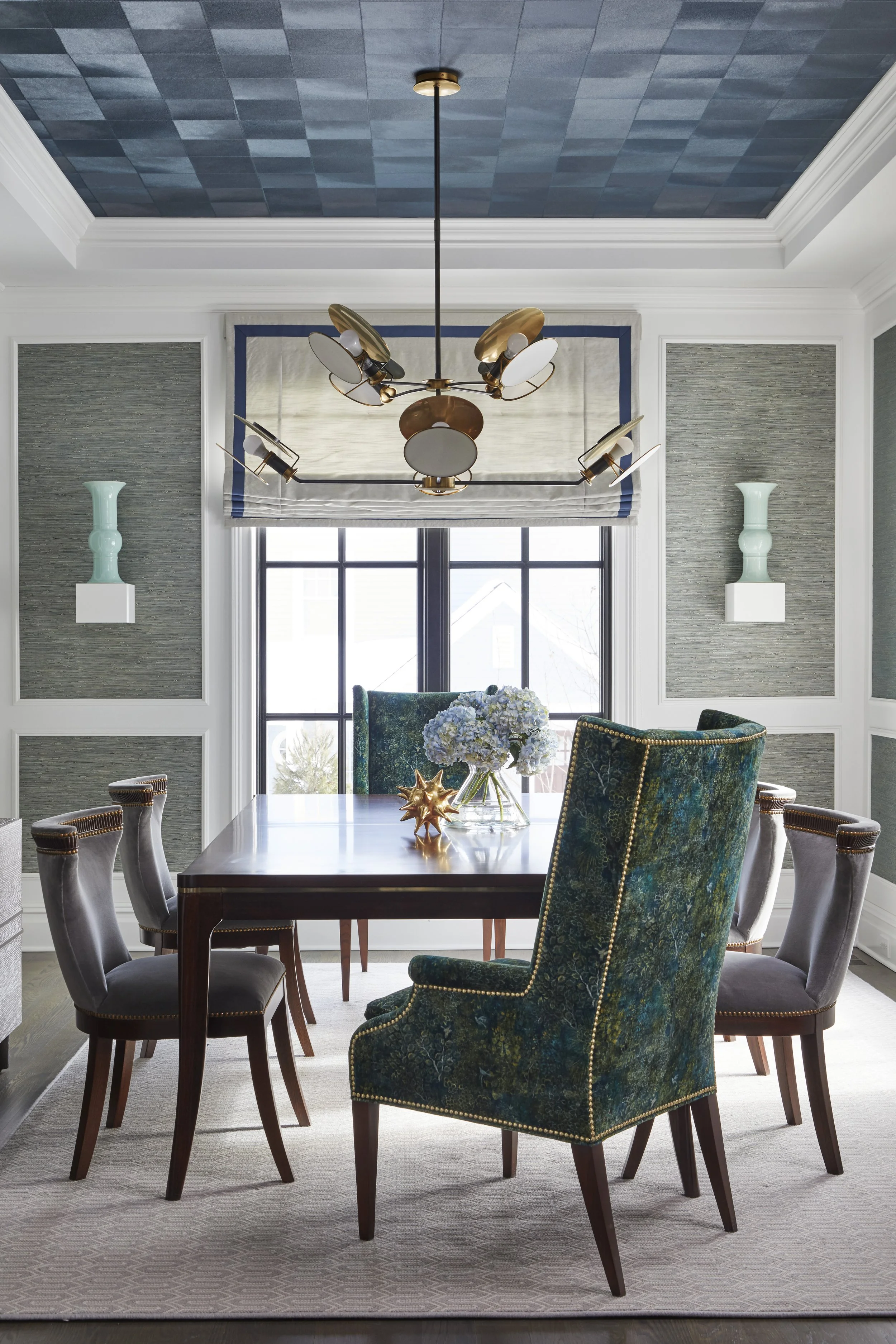 A dining room with a dark wood table surrounded by a mix of velvet upholstered chairs and wooden chairs, a floral centerpiece on the table, a large window with white framing, textured wallpaper, wall-mounted turquoise vases, and a gold and black chan