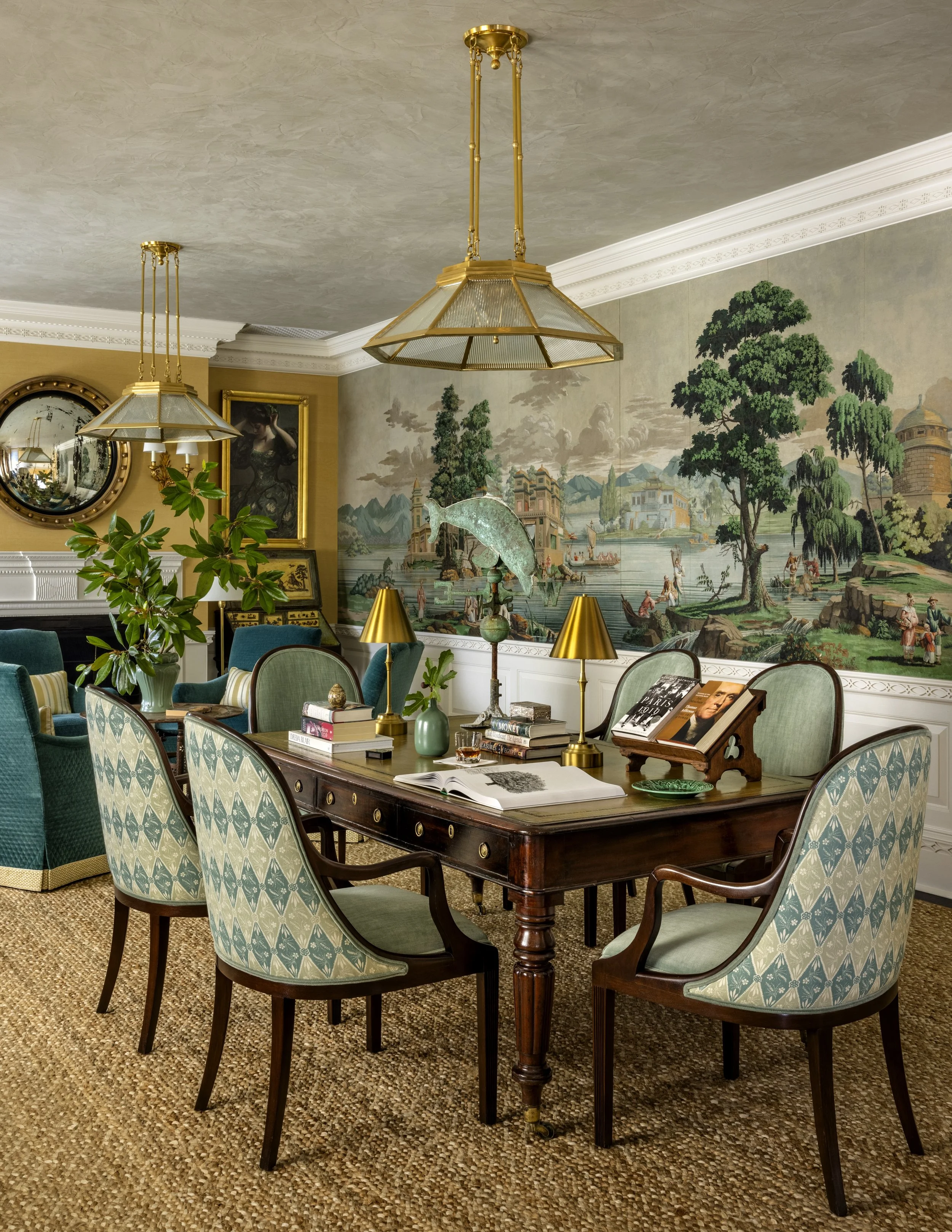 A dining room with a large wooden table surrounded by upholstered chairs with patterned fabric. The table has stacks of books, a small green vase, and a wooden display stand. There are gold table lamps, a large mirror, and artwork on the wall behind.