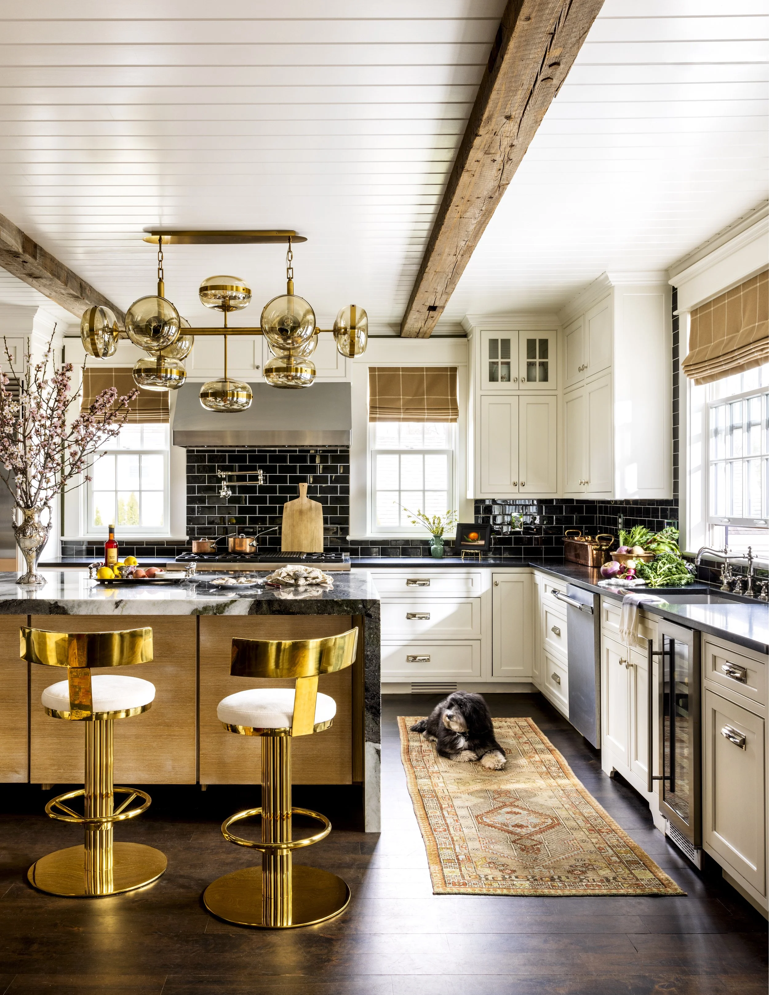 View of a modern kitchen with white cabinets, black backsplash, and large windows. There is a kitchen island with a marble countertop, paused at the edge of the image. Two gold barstools with cream cushions are placed at the island. A black dog is ly