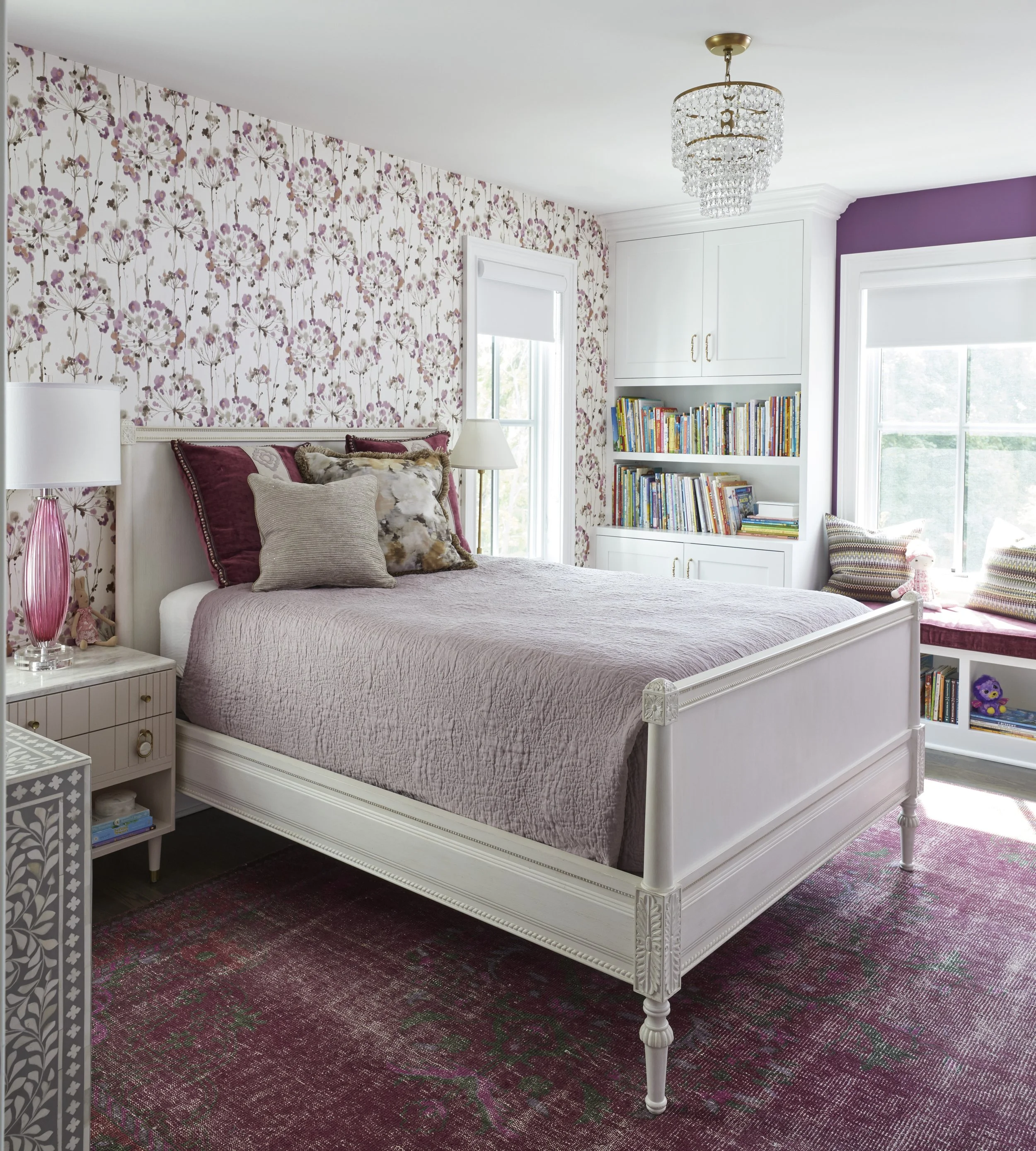 A bedroom with floral wallpaper on one wall, a white bed with multiple pillows, including a furry one, and a pink lamp on a nightstand. There are bookshelves filled with colorful books and a window with white blinds, a purple accent wall, and a chand