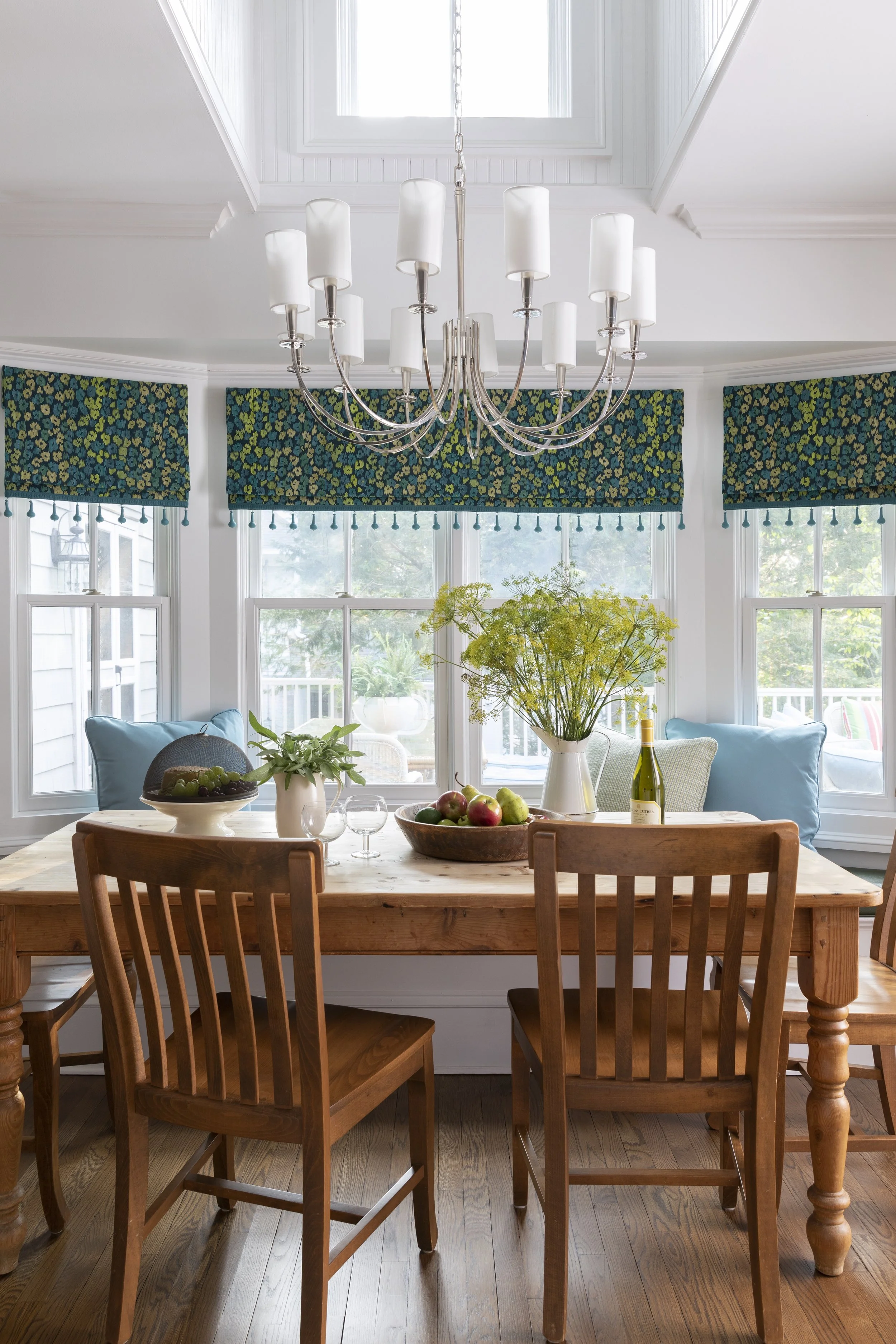 Dining room with a wooden table, four matching wooden chairs, a large bowl of apples, a white vase with yellow flowers, a bottle of wine, two glasses, and other small plants. Behind the table are large windows with patterned blue and green valances, 