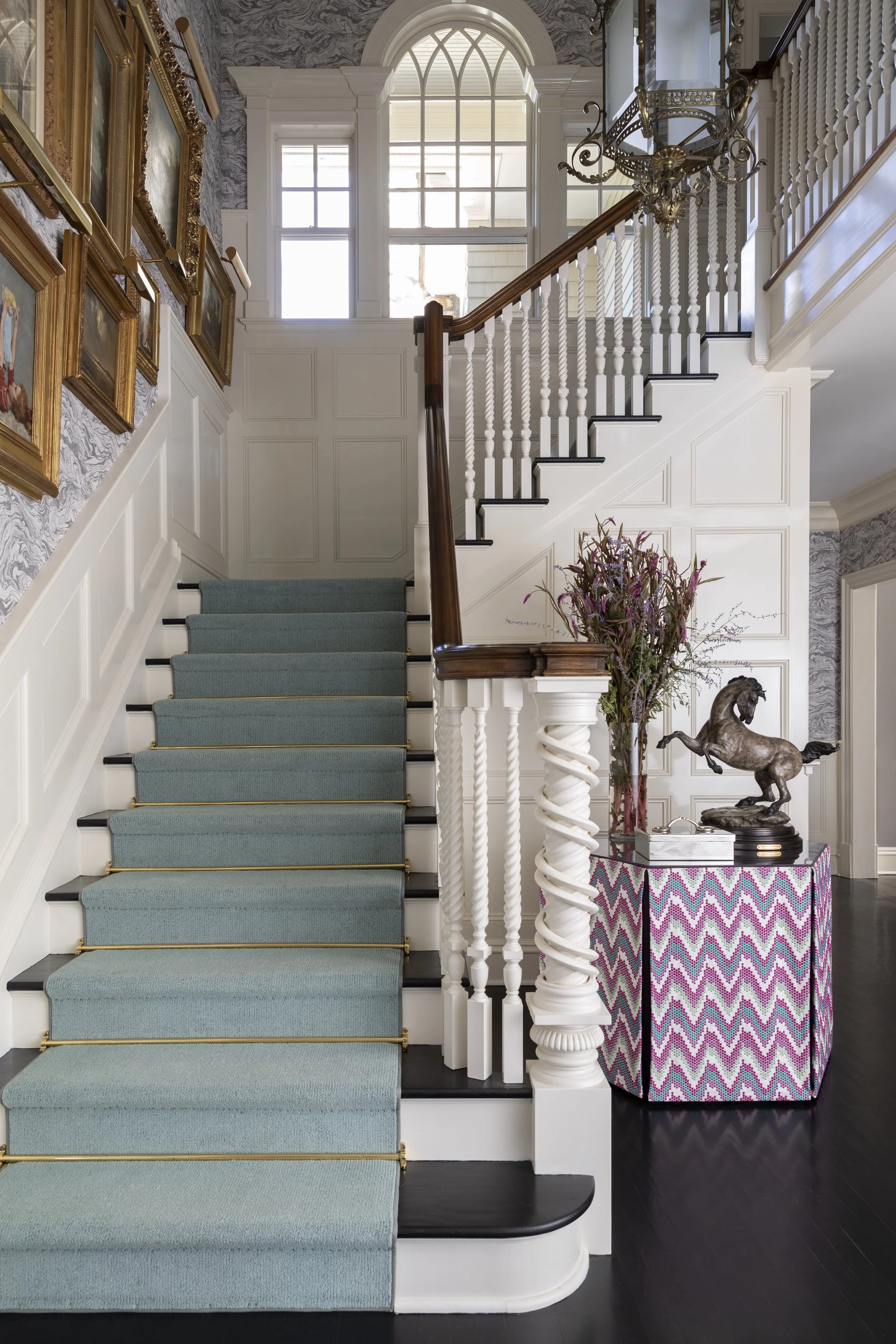 A staircase with white balusters, wooden handrail, and teal carpeted steps, next to a wall with framed paintings. Large windows above illuminate the space, and there is a decorative table with a colorful chevron pattern, a statue of a horse, and a bo