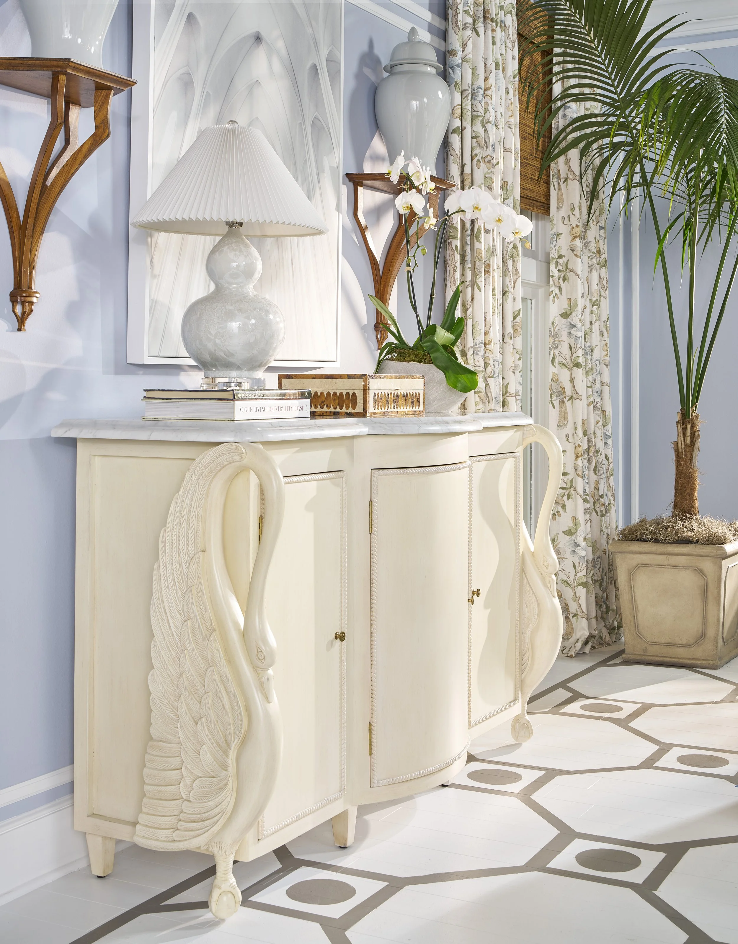 An interior corner of a room featuring a white vintage-style cabinet with ornate carved swan legs, a marble top, and beige panels. On top of the cabinet are a stack of books, a large white ceramic table lamp with a pleated shade, a decorative wooden 