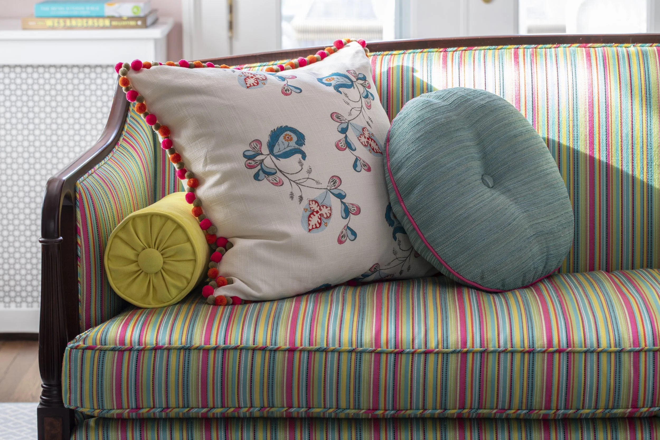A colorful striped sofa with decorative pillows including a rectangular pillow with embroidered flowers and pom-pom trim, a yellow cylindrical bolster, and a large round teal pillow with pink piping.