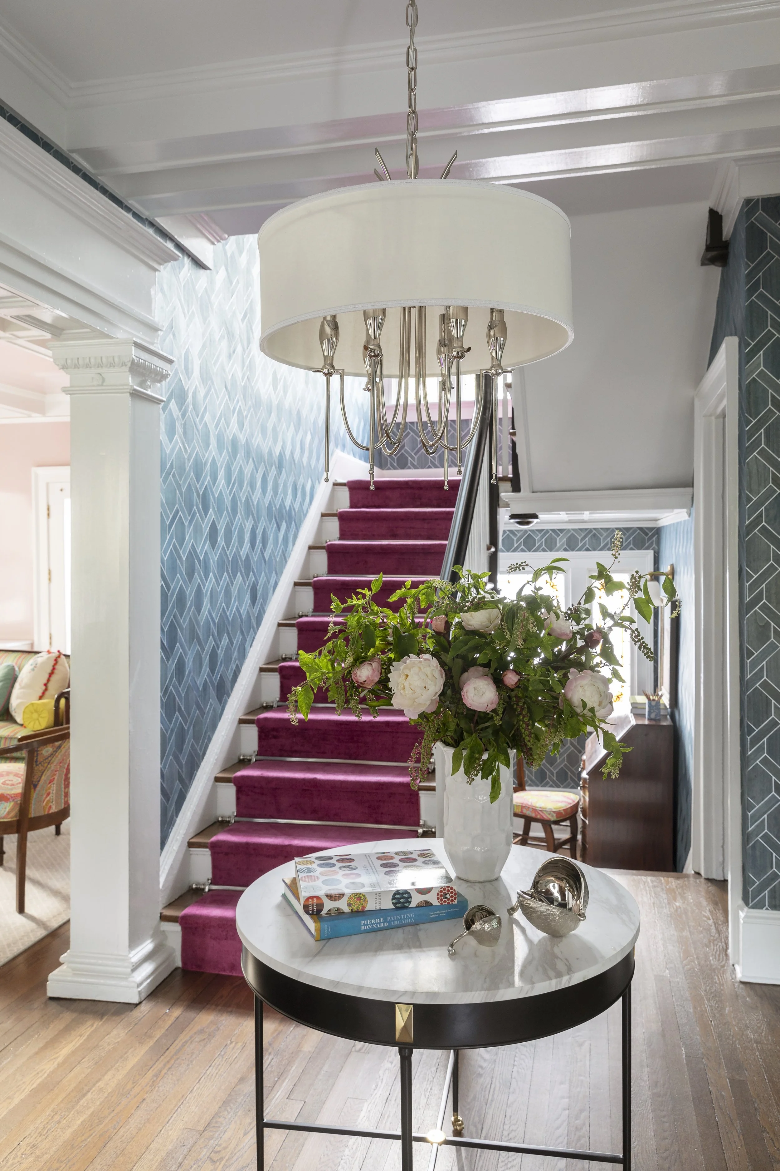 Interior of a house with a staircase covered in a pink carpet, blue patterned wallpaper, a white column, a lady with a plant on a table, and decorative items including books and silver objects.