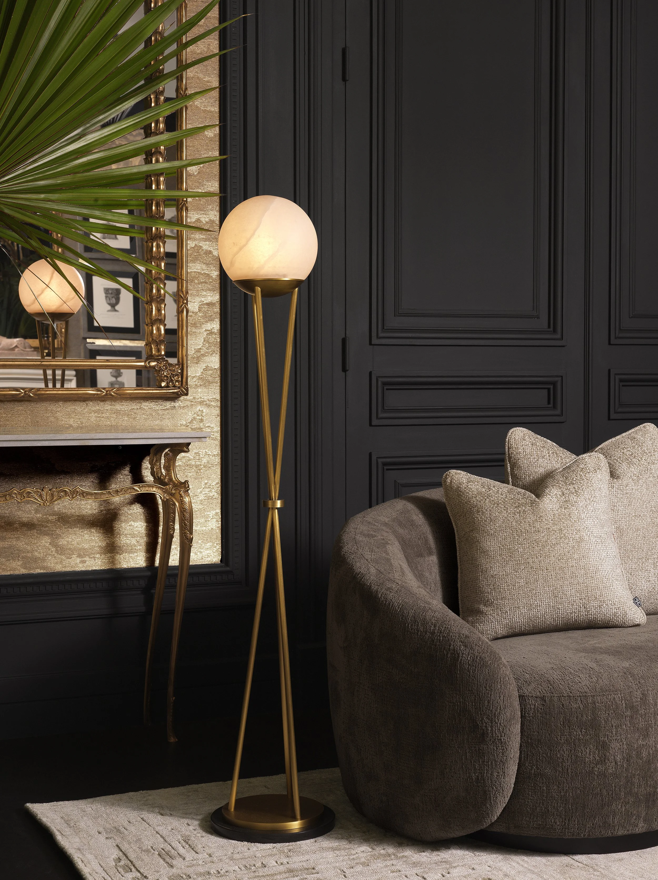 A living room corner with a black paneled wall, a beige armchair with pillows, a gold floor lamp with a marble globe shade, a gold-framed mirror, and a gold console table with framed pictures and a large green plant.