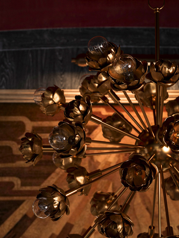 Close-up of a modern chandelier with multiple bronze-colored flower-shaped fixtures and exposed light bulbs, hanging in a room with dark walls and wooden flooring.