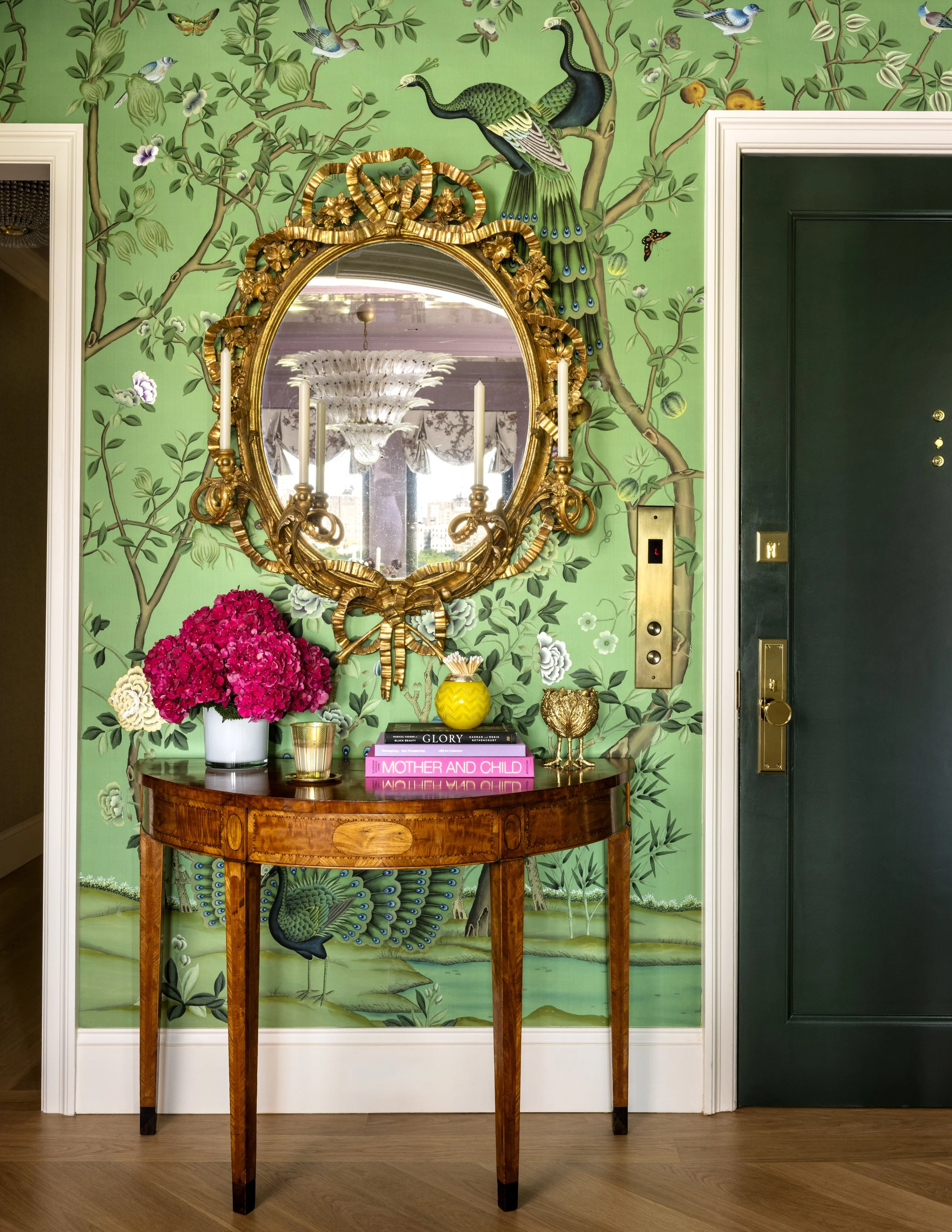 A gold-framed mirror hangs on a green wall decorated with a mural of peacocks, birds, and floral designs. A wooden table below the mirror holds a pink hydrangea flower arrangement, books, and decorative objects. An elevator door is visible to the rig