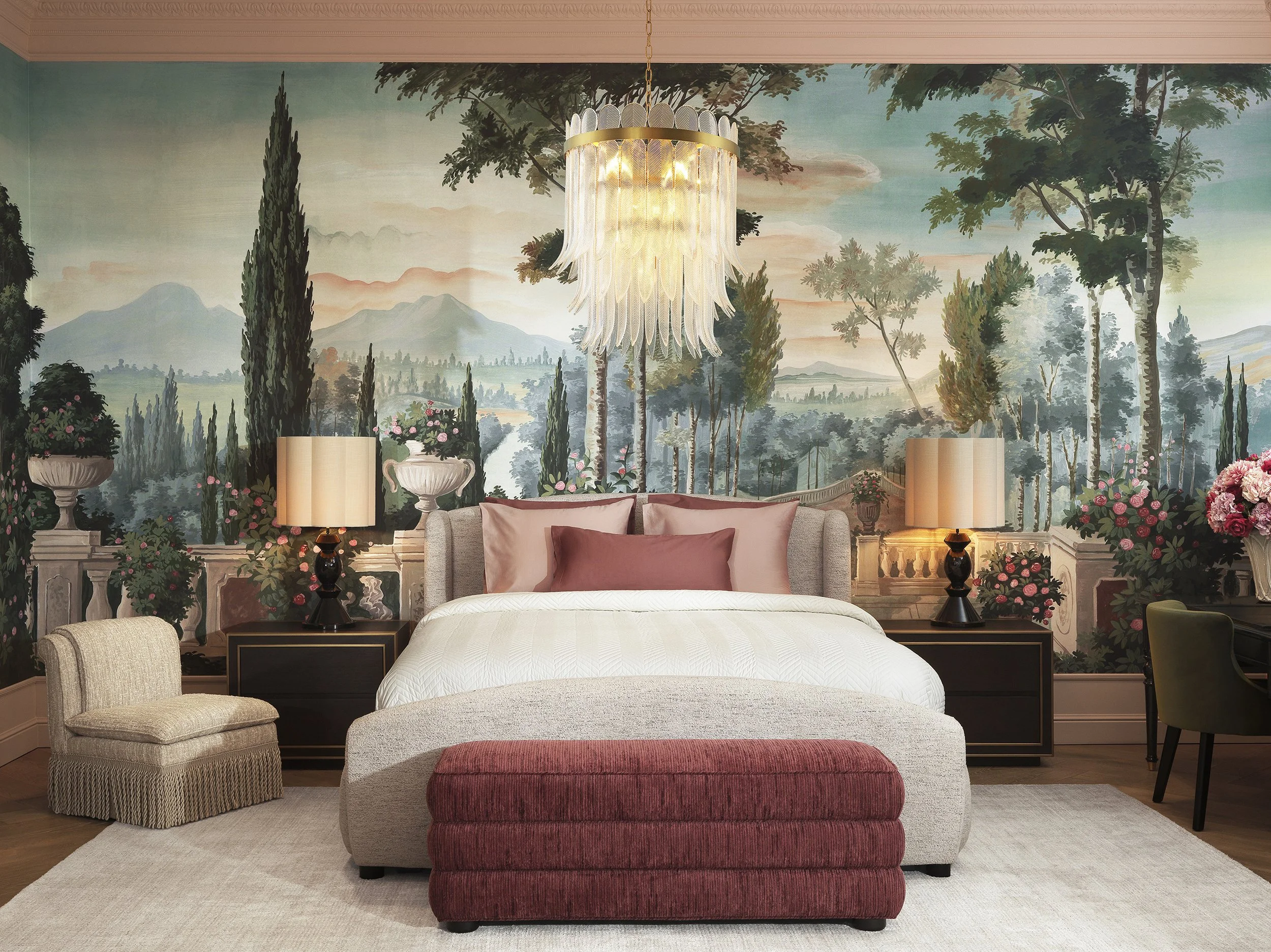 A bedroom with a mural of a landscape featuring mountains, trees, and a sunset sky. There is a hanging chandelier, a bed with pink pillows, two lamps on bedside tables, an armchair, a cushioned ottoman, and various potted plants with pink flowers.