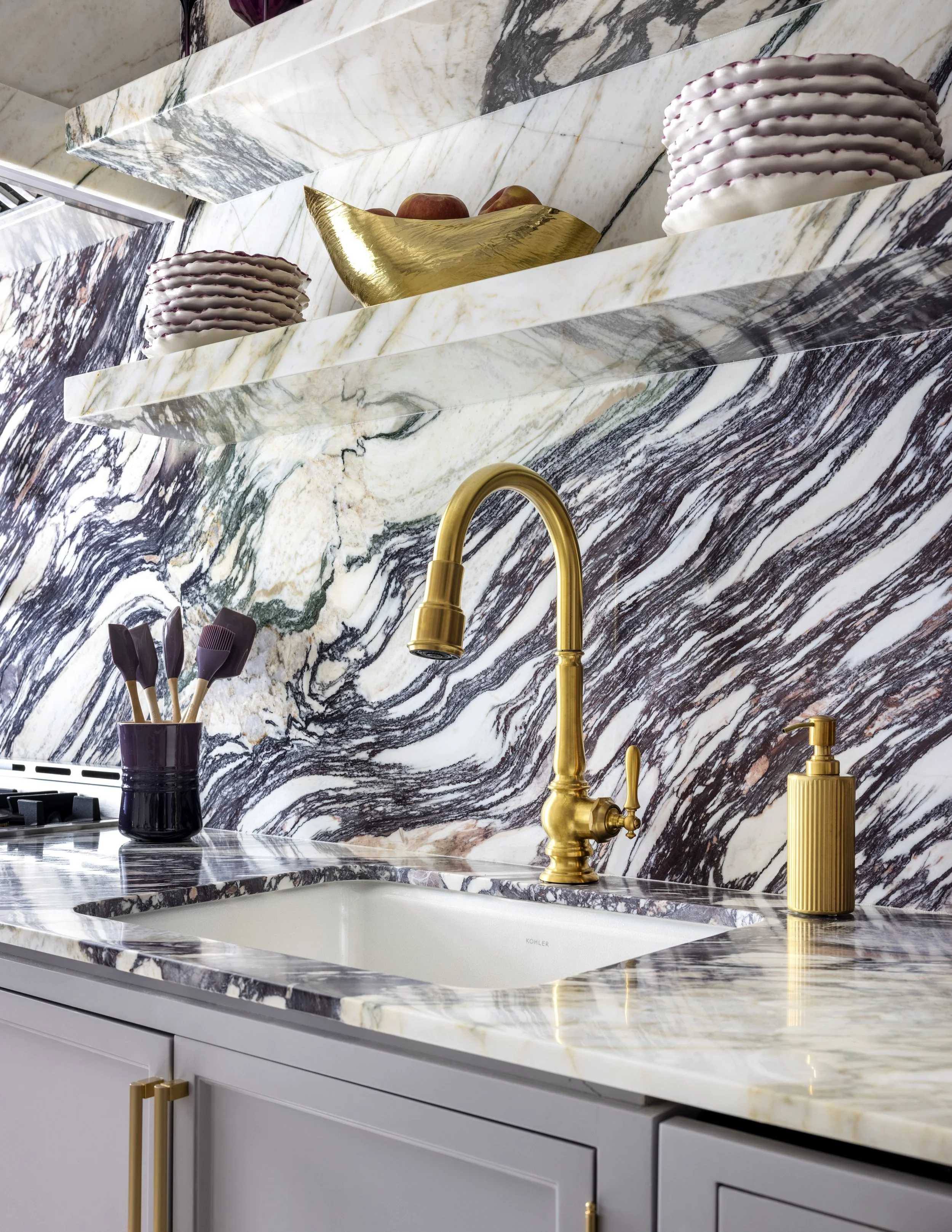 A kitchen sink area with a white and black marble backsplash and countertop, gold faucet, and gold hardware on the light gray cabinetry. Decorative items include a dark purple container with kitchen utensils, a golden bowl with apples, and a stack of
