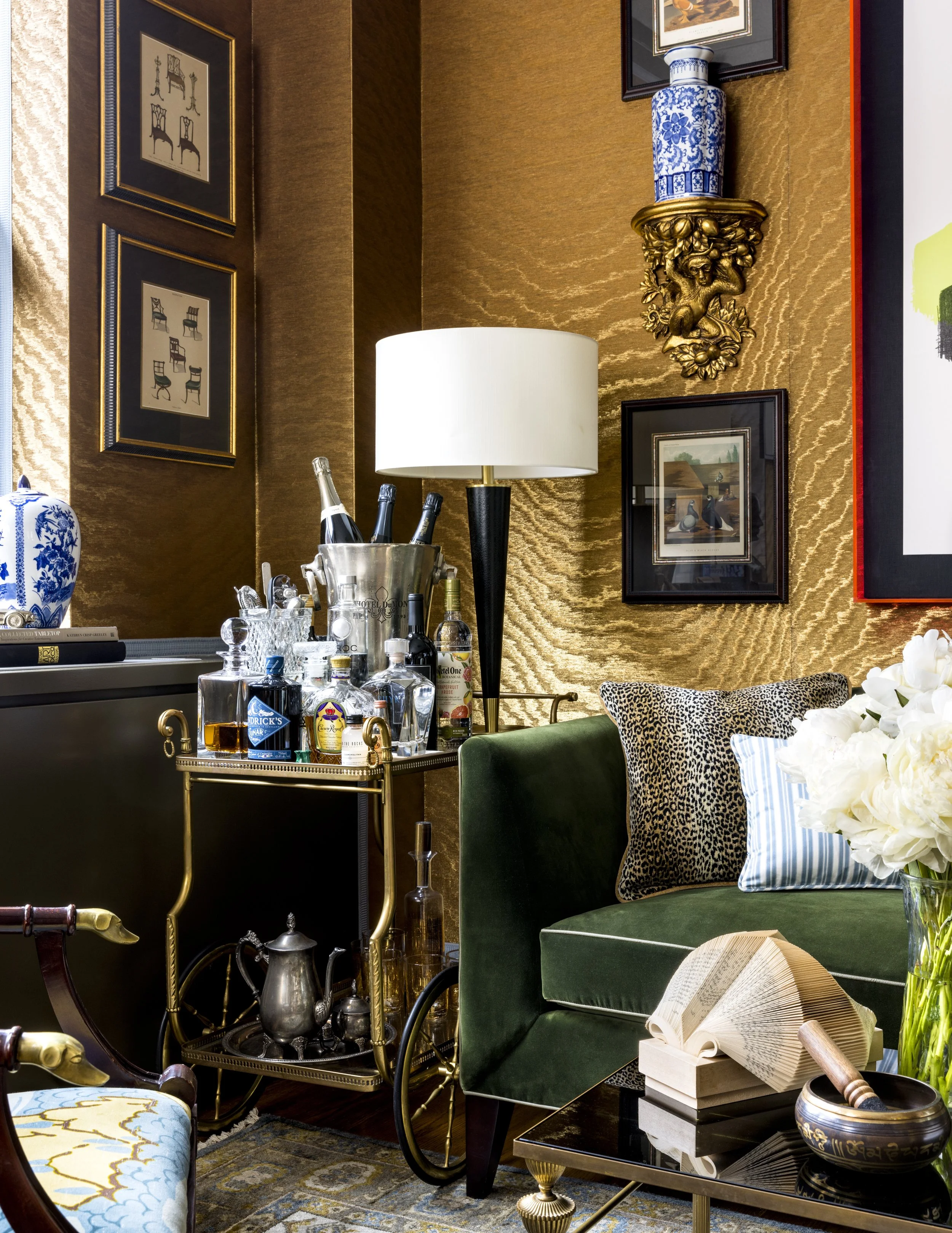 Interior of an elegant room with gold textured walls decorated with framed artwork and a blue and white ceramic vase on a gold ornate wall mount. There is a white lampshade on a black and gold lamp, a green velvet chair with patterned pillows, a smal