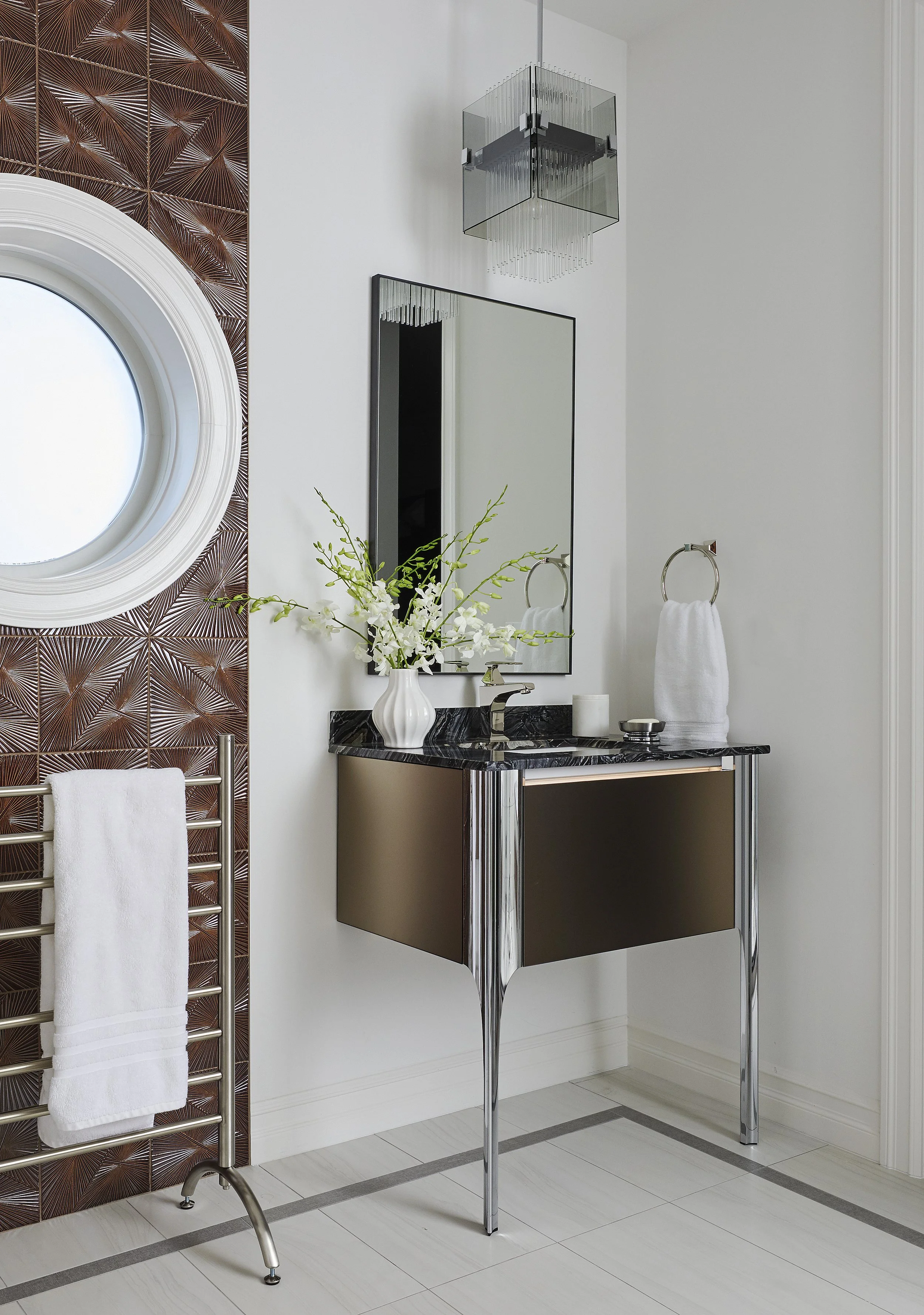 Modern bathroom with a floating vanity with a black marble top, a faucet, and a mirror. A tall vase with white flowers decorates the vanity. To the right, a silver towel ring holds a white towel. On the left, a metallic towel rack has a white towel. 