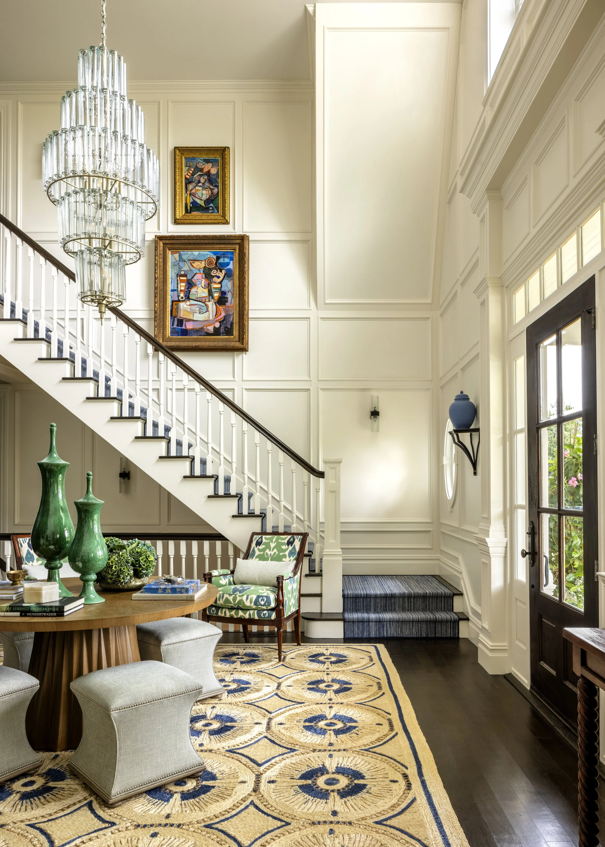 Living room with white walls, staircase, gold-framed colorful abstract paintings, large glass chandelier, green decorative vases, patterned armchair with blue and green fabric, beige patterned area rug, dark wood flooring, entrance door with glass pa