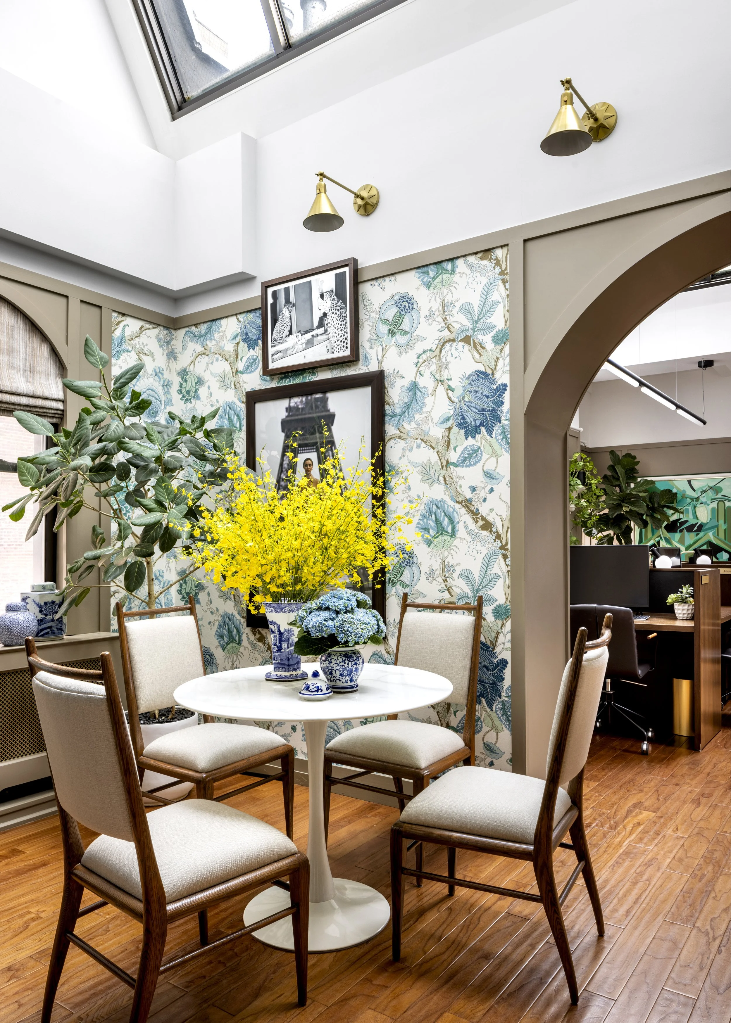 A dining area with a white round table surrounded by six upholstered chairs with wooden legs. The table has blue and white china vases with yellow and blue flowers. There's a large potted plant with broad leaves and wallpaper with a blue floral patte