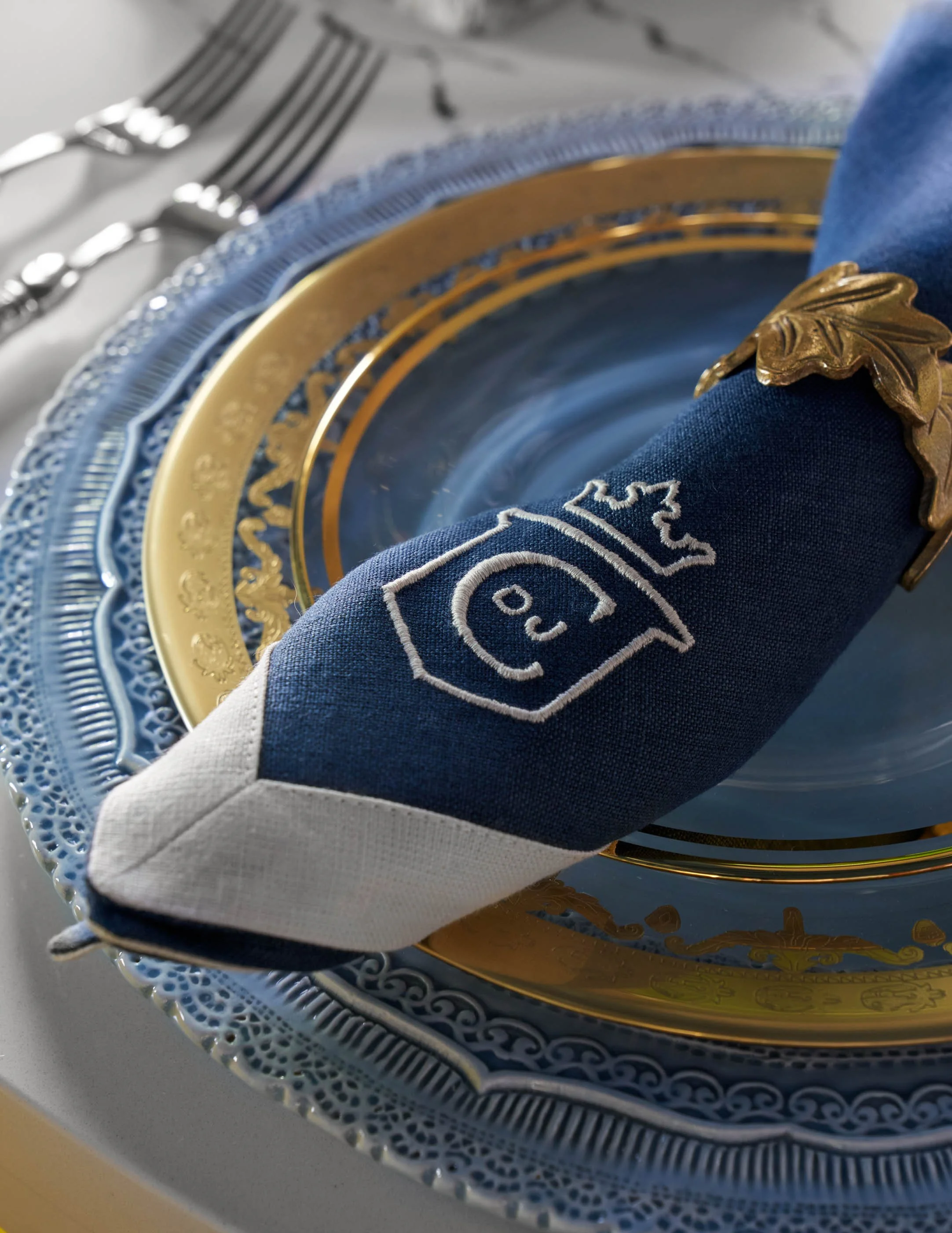 Close-up of a decorative plate with a gold rim, a dragon emblem, and a navy blue cloth napkin with embroidered crest, accompanied by silver forks on a table.