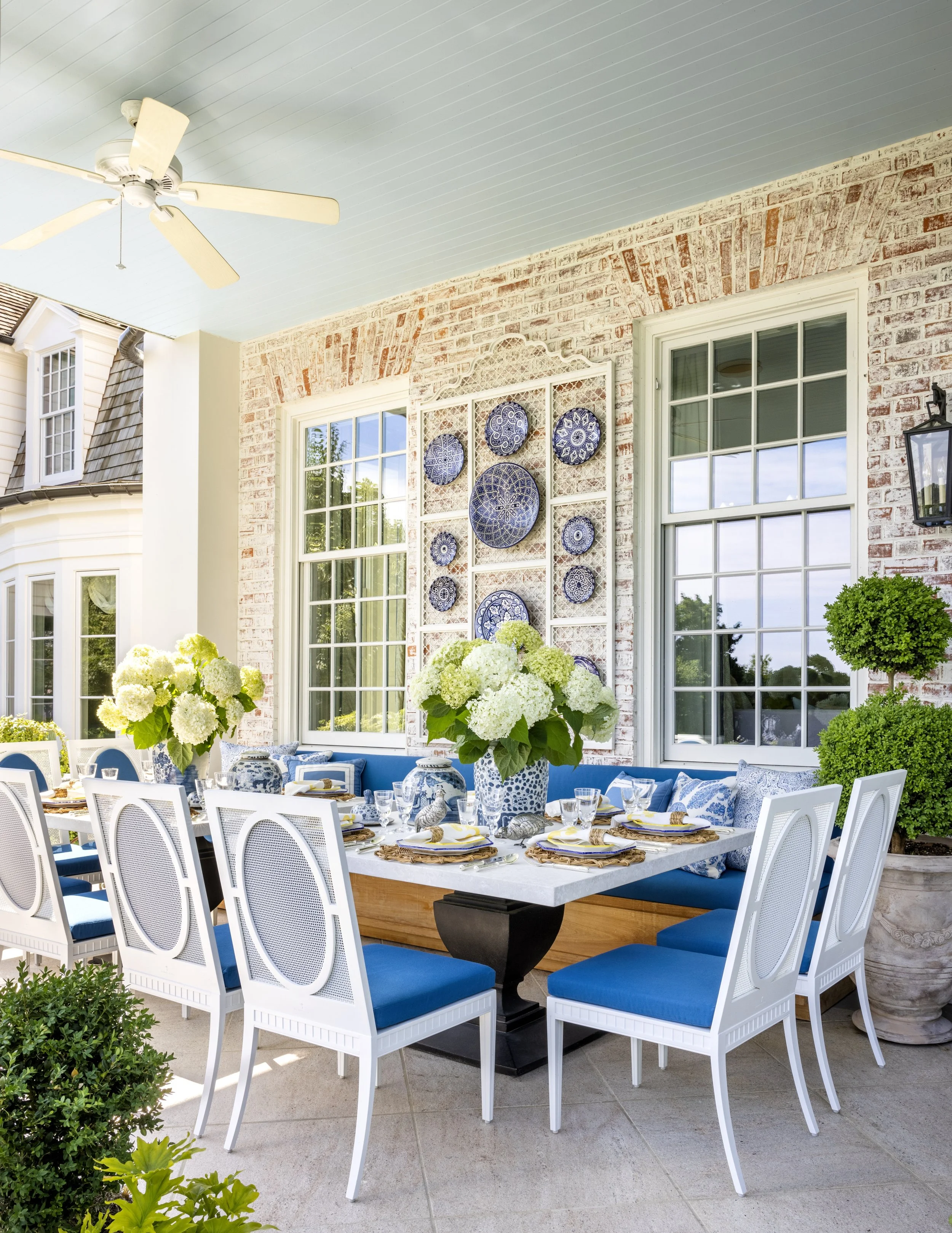 Outdoor patio with dining table set for a meal, decorated with white and blue plates, hydrangea flowers, and surrounded by white chairs with blue cushions, featuring large windows, brick wall, and decorative plates art, with outdoor plants, under a b