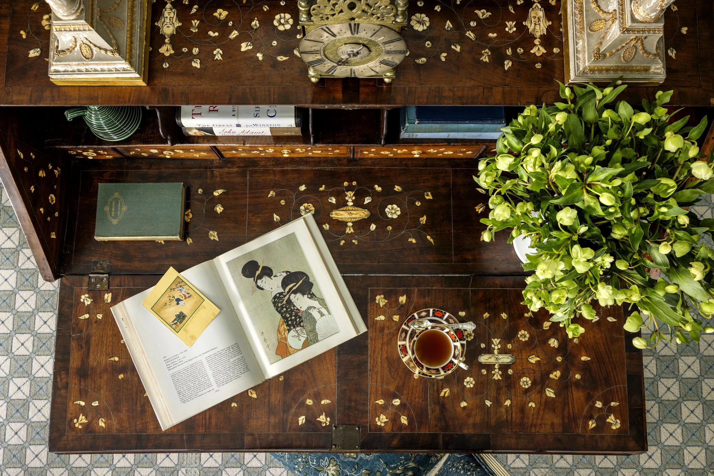 A wooden table with gold floral inlay, displaying an open book about Japanese art, a teacup filled with tea, a green book titled "Deception," a closed blue book, a chessboard, and shelves with books and decorative items. A large green plant with budd